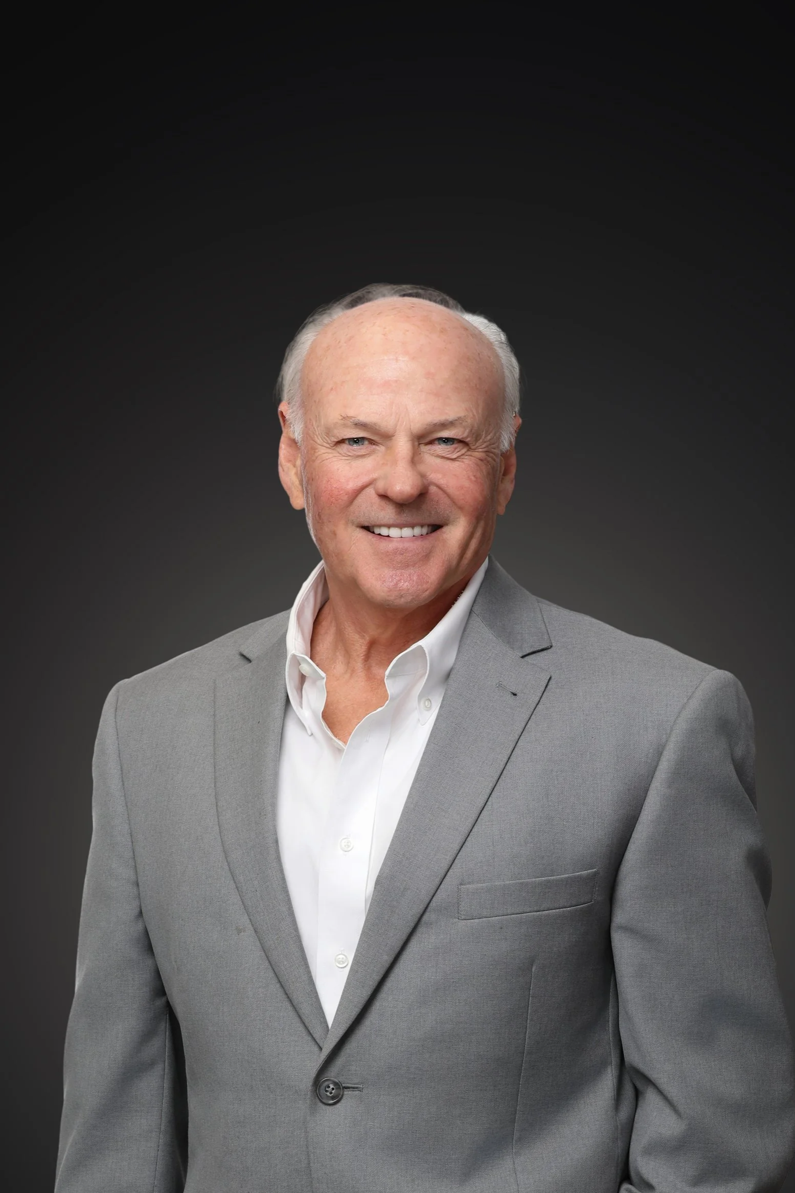 Close-up portrait of a smiling man wearing a black tuxedo, white shirt, and black patterned tie, against a dark background.