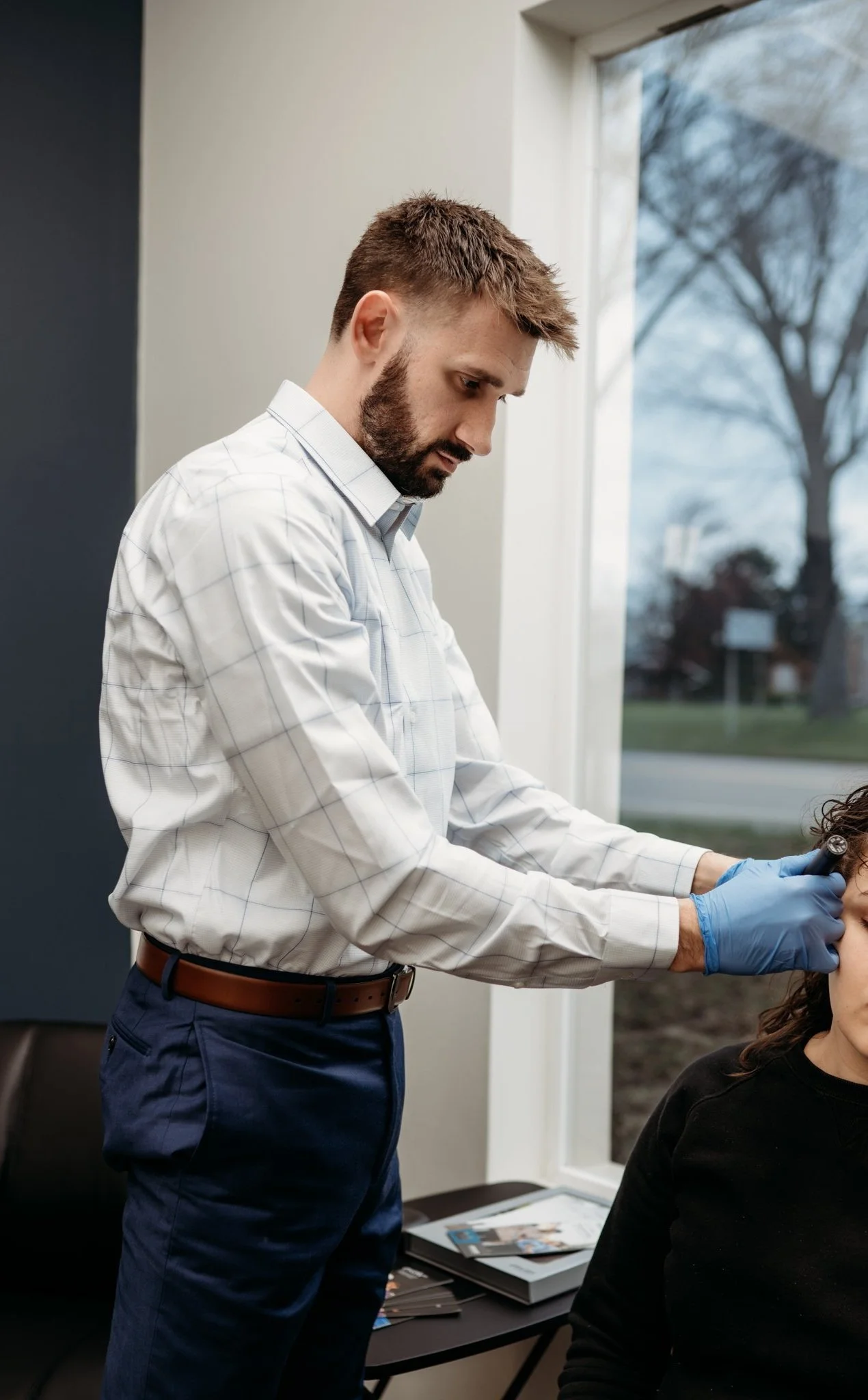 A man with a beard and short hair, wearing a checkered shirt and blue pants, is getting a tattoo on his head by a tattoo artist in a room with large windows showing trees and a street outside.