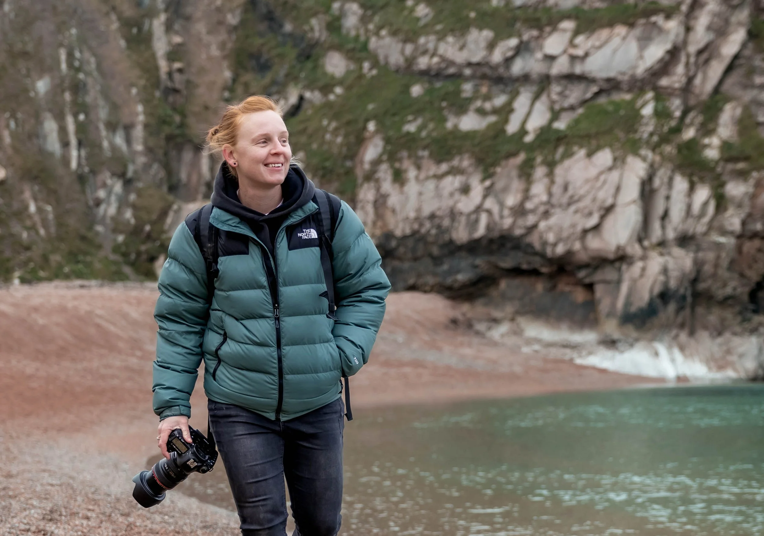 A woman with red hair, wearing a teal puffer jacket and black jeans, walking along a lakeshore while holding a camera in her right hand, with rocky cliffs and trees in the background.