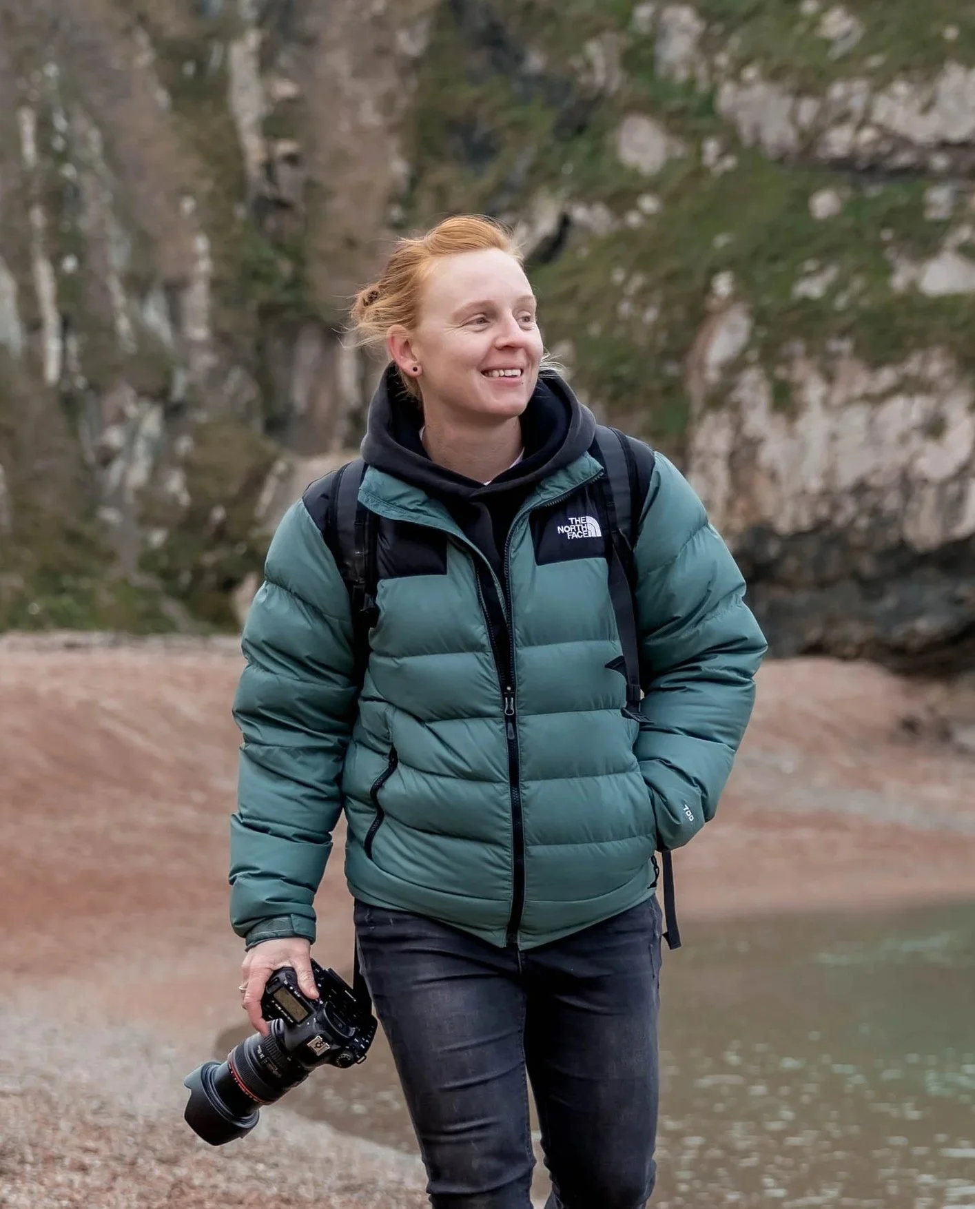 A woman with red hair wearing a green puffer jacket and black pants, holding a camera, and walking outdoors near a body of water with rocky and tree-covered surroundings.