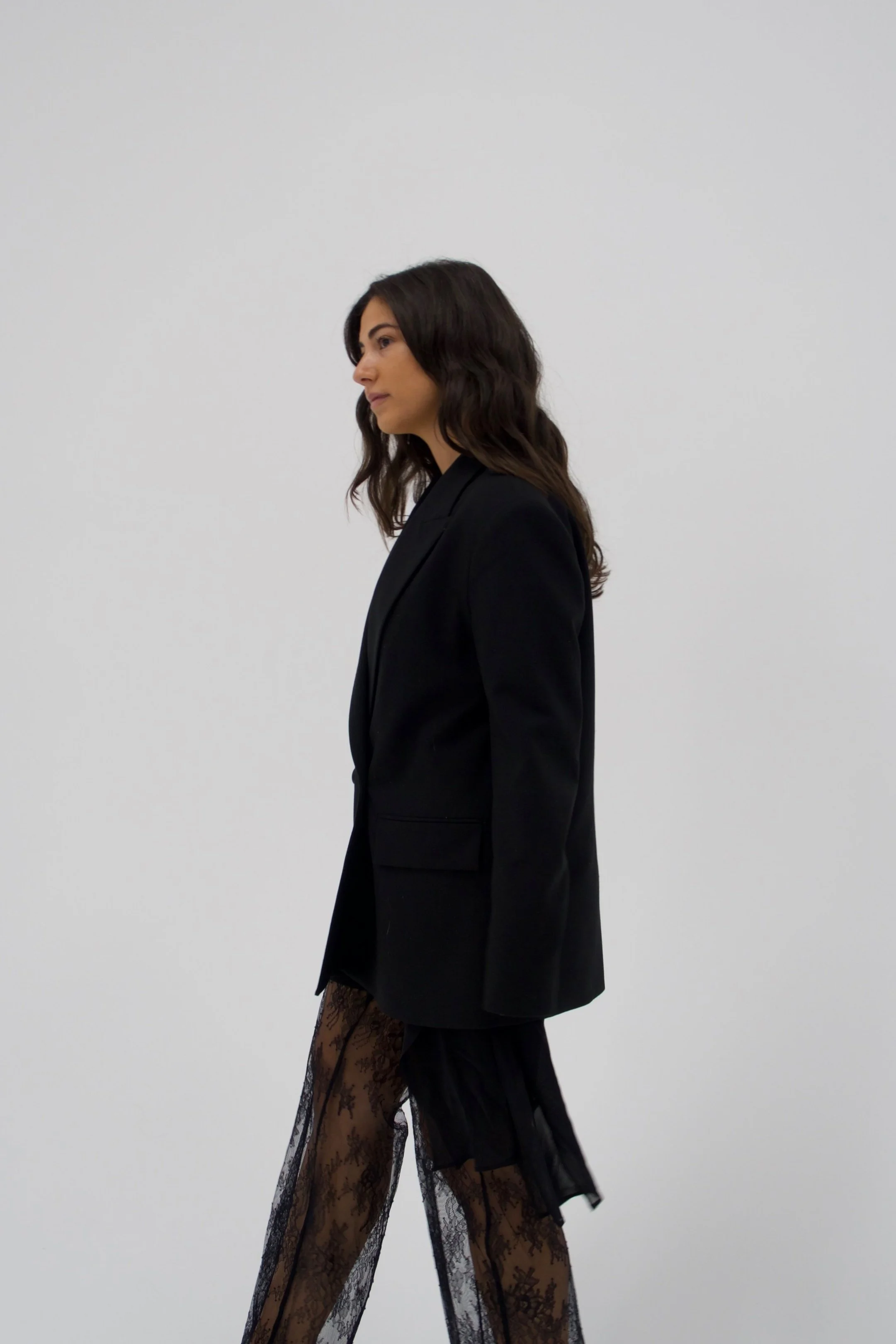 Side view of a woman in a black blazer and sheer black lace pants standing against a plain white background.