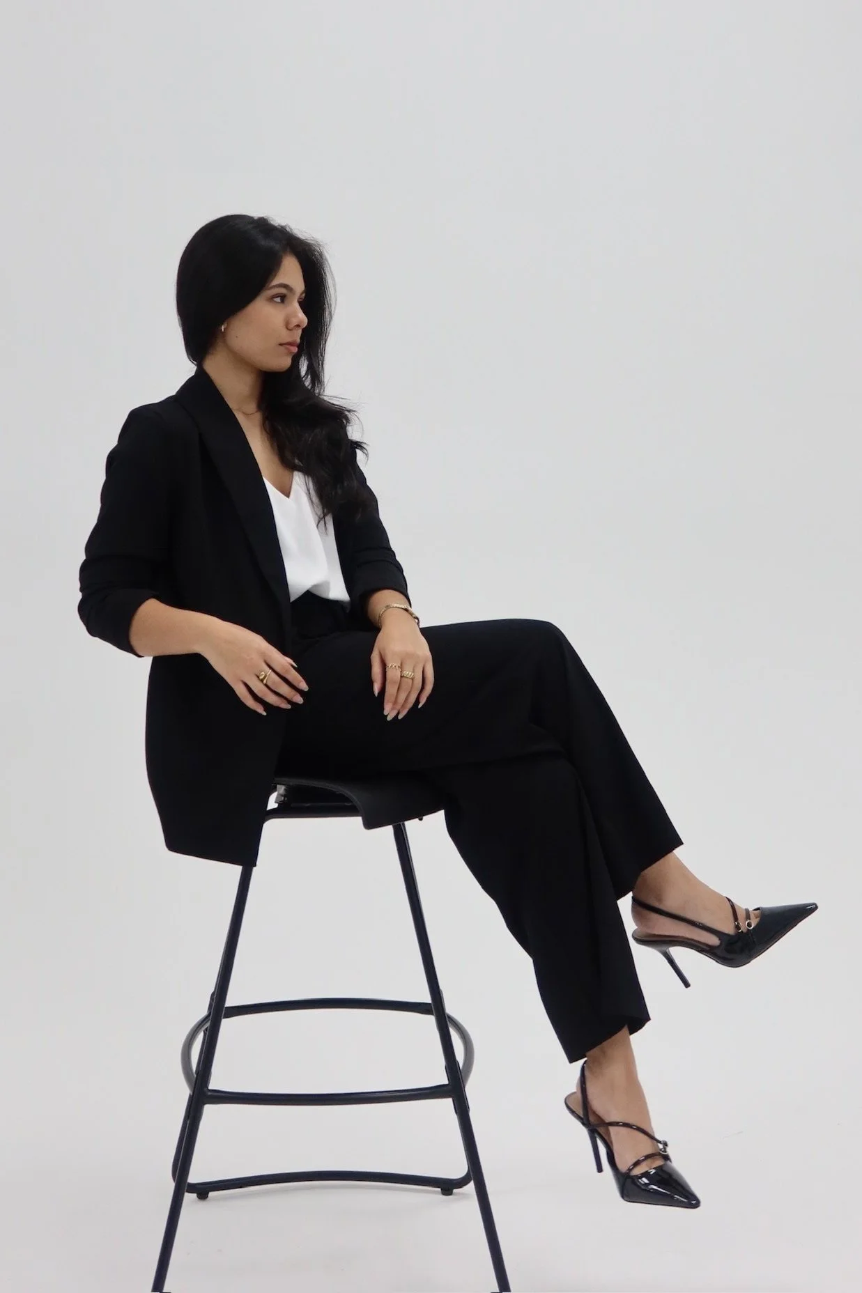 A woman in a black blazer, black pants, and high heels sitting on a tall black stool against a plain white background.