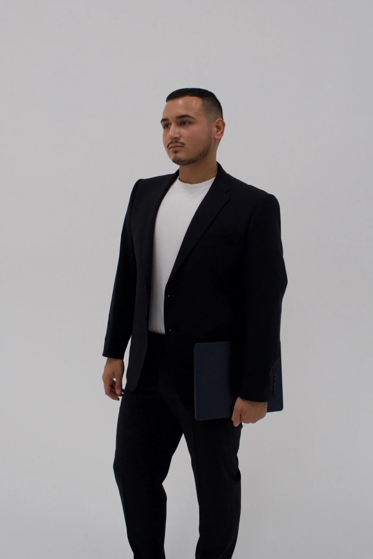 A man in a black suit holding a tablet standing against a plain white wall.