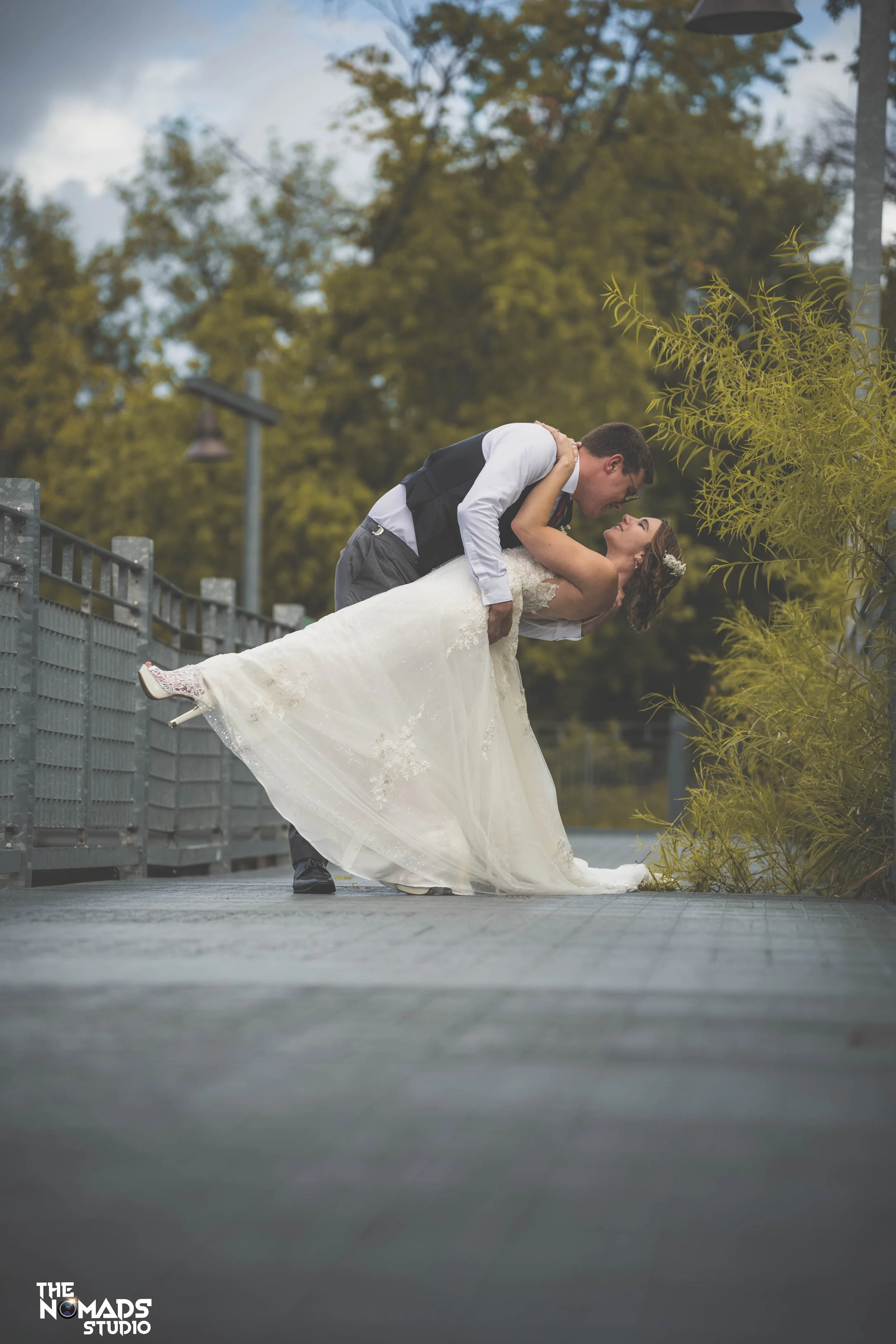 A bride and groom in wedding attire are on an outdoor deck, with the groom dipping and kissing the bride. The bride is smiling, wearing a white lace gown, and the groom is wearing a dark vest and glasses. There are green trees and a cloudy sky in the