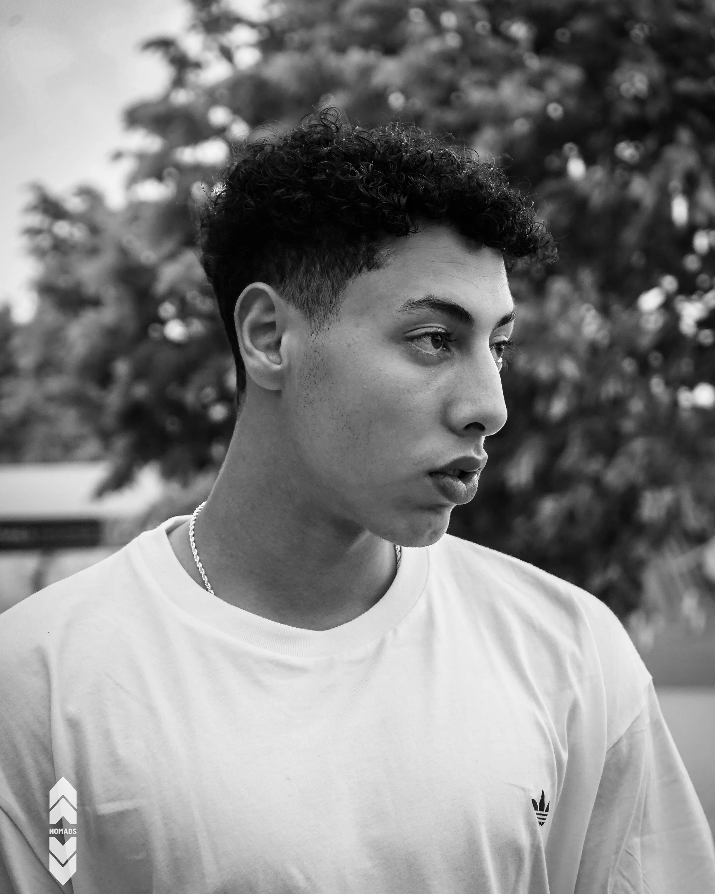Close-up of a person's face with short curly hair, wearing a white t-shirt with an Adidas logo and a silver chain, outdoors with blurred trees in the background.