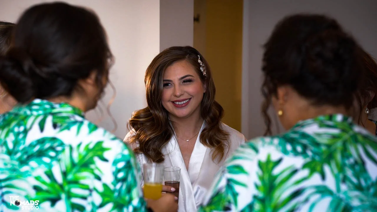 A woman with brown hair and a white satin robe smiling at two women in green and white leaf-patterned shirts during a social gathering, each holding a drink.
