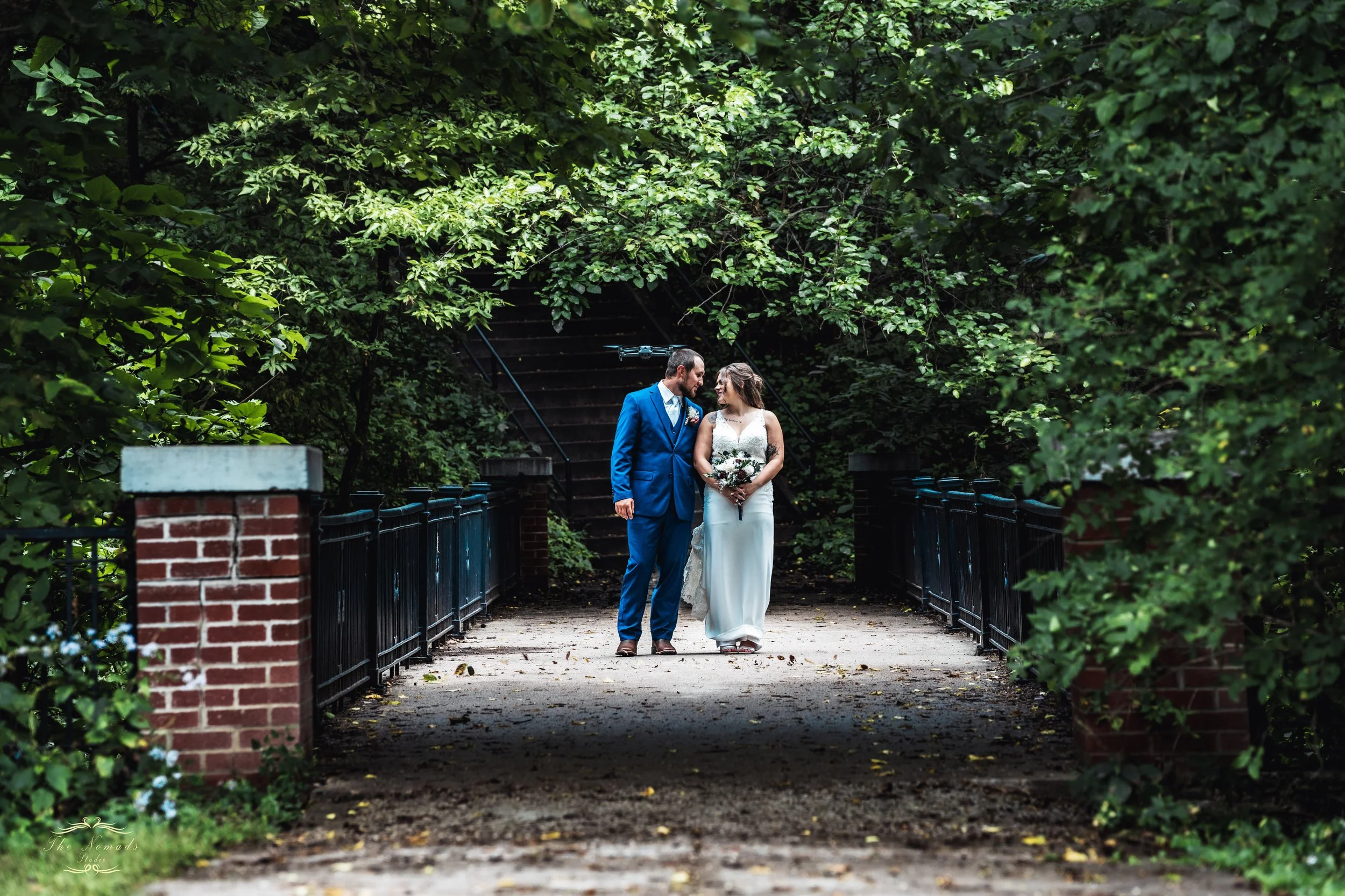 A bride and groom walking hand in hand on a park pathway surrounded by lush green trees, with stairs and black fences on each side.
