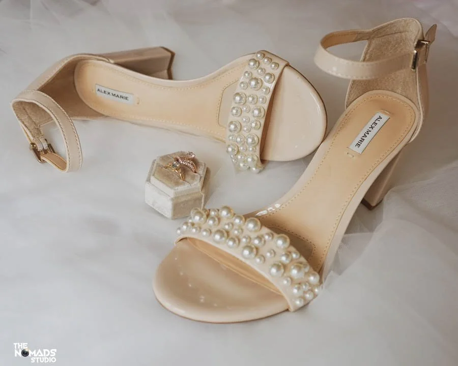 A pair of beige high-heeled shoes with ankle straps and pearl embellishments on the front strap, along with a ring in a small box.