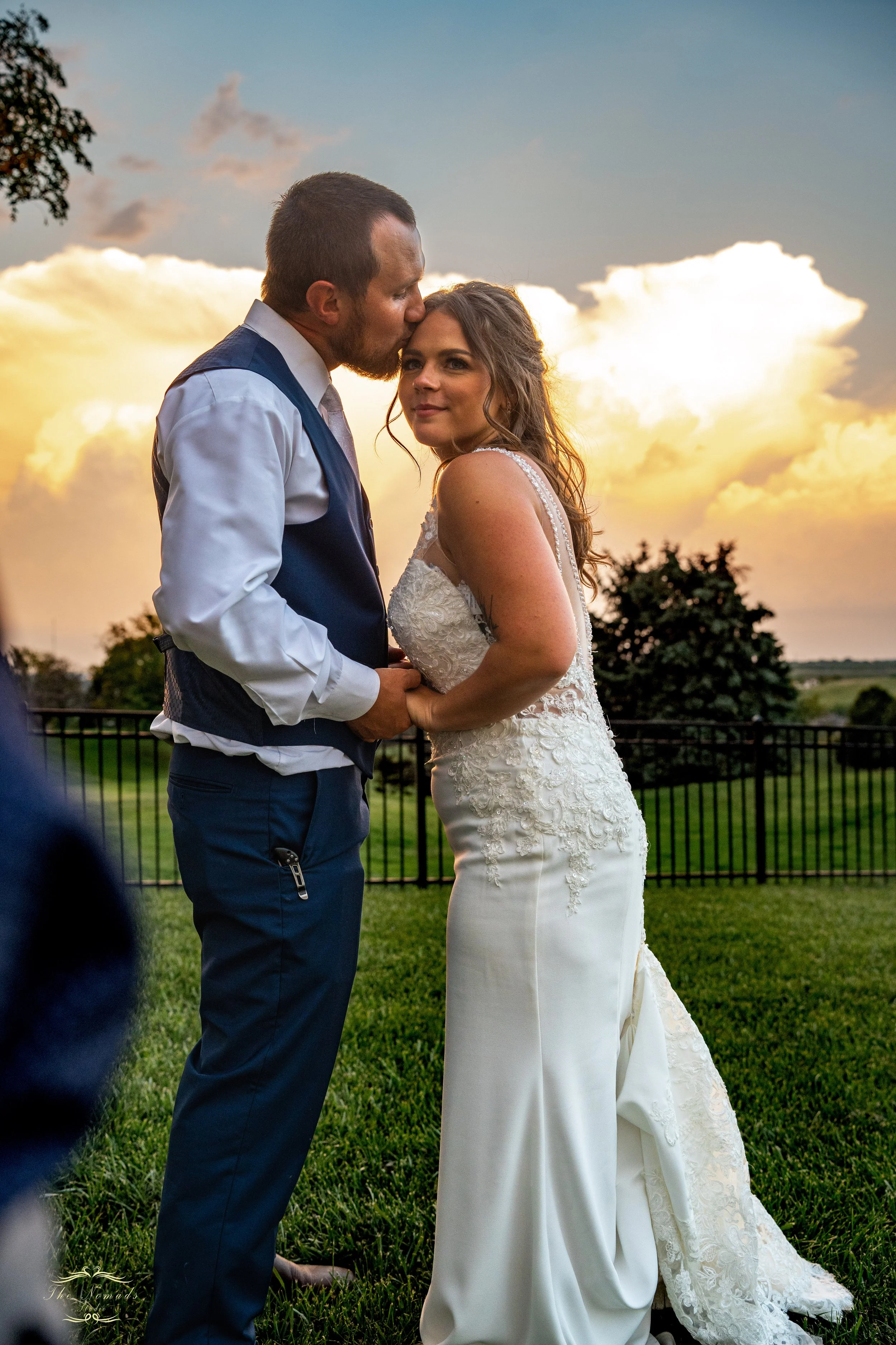 A bride and groom standing outdoors at sunset, holding hands, with the groom kissing the bride's forehead, and trees and a fence in the background.