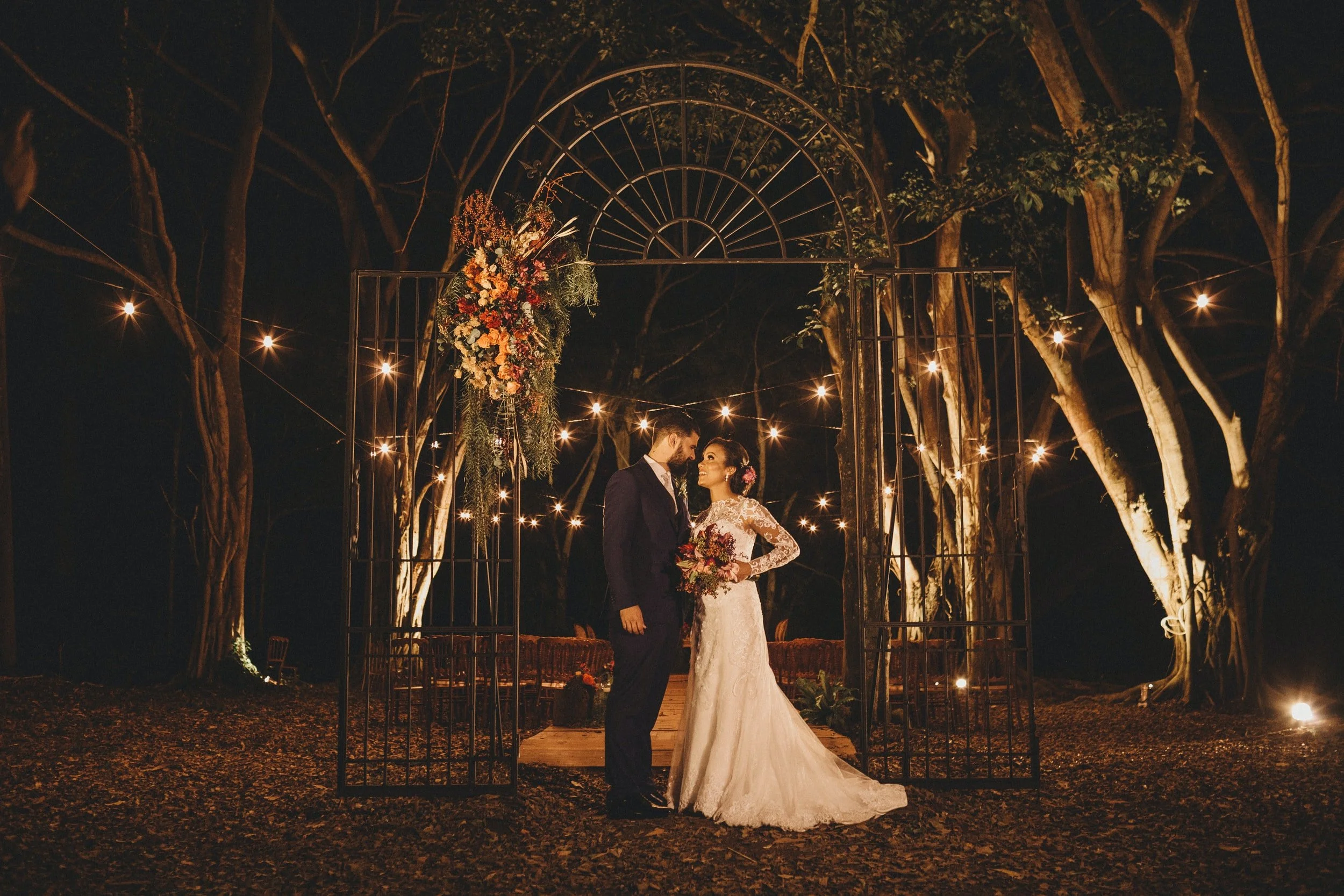 A bride and groom standing at an outdoor wedding altar decorated with flowers and string lights at night, with trees in the background.