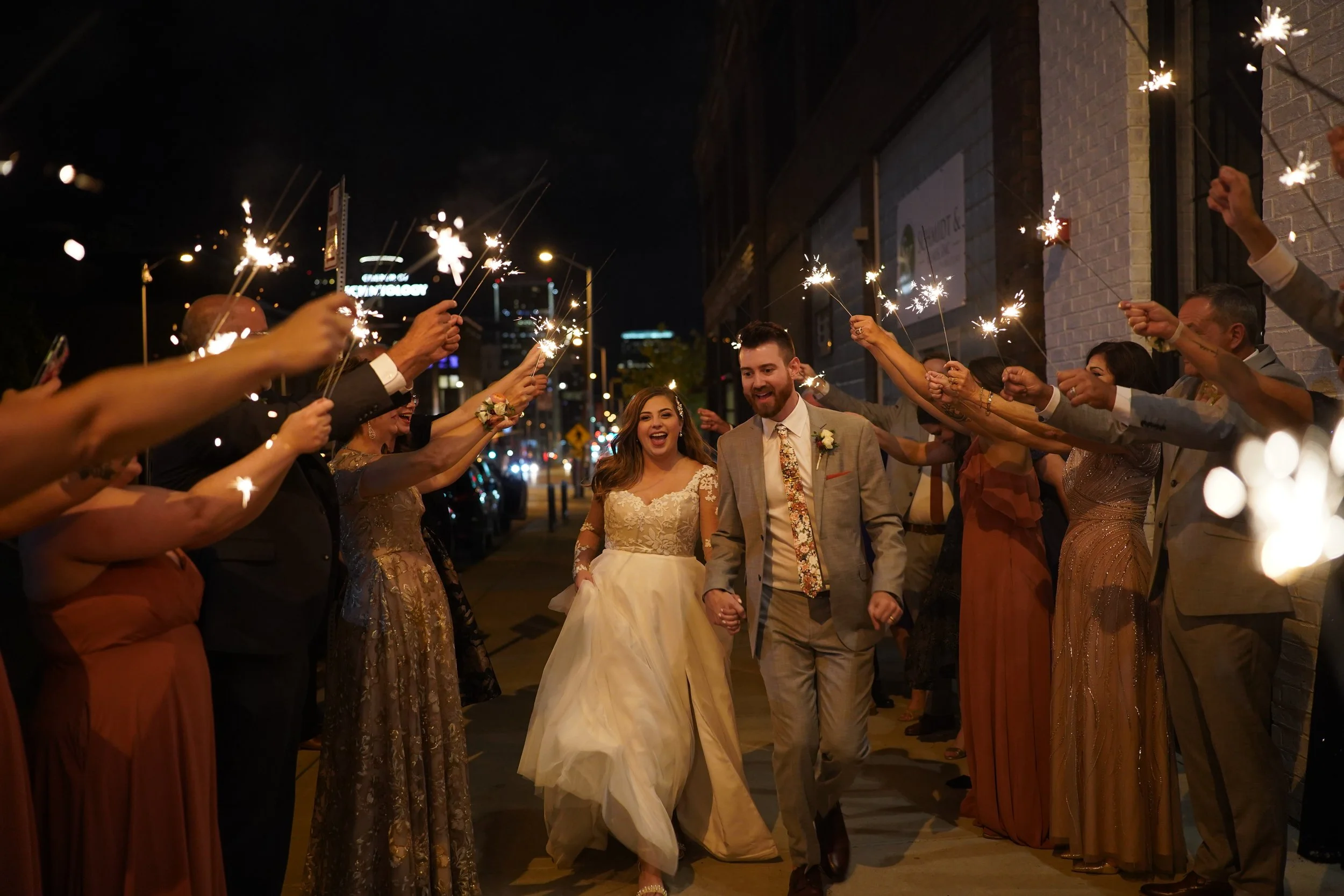 A group of people holding sparklers forming a pathway for a newlywed couple to walk through at night in an urban setting.