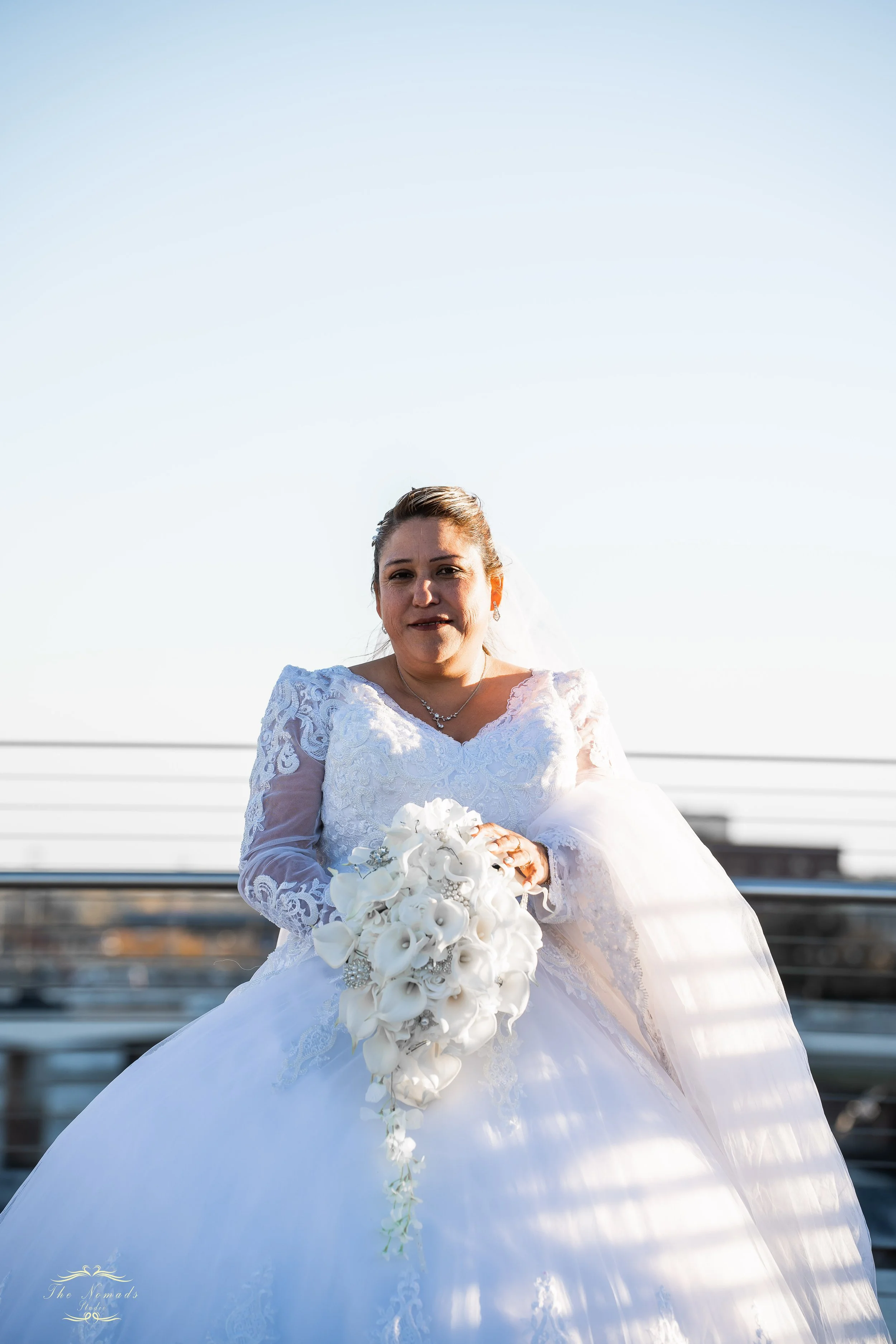 A bride in a white wedding dress holding a bouquet of white flowers, standing outdoors on a sunny day with a clear blue sky in the background.