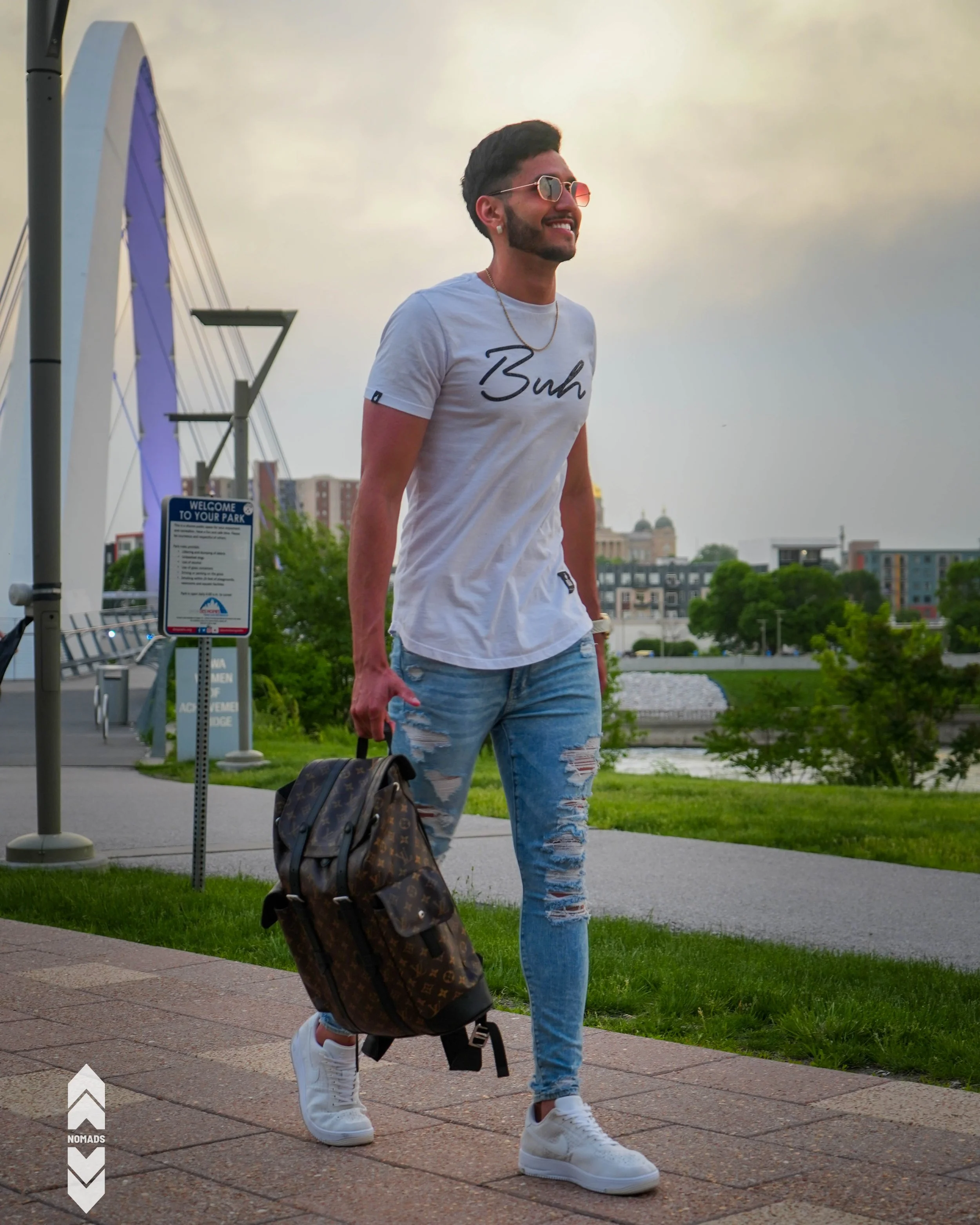 A man walking outdoors near a park with an urban background, wearing sunglasses, a white t-shirt that says 'Buh', ripped jeans, and white sneakers, carrying a Louis Vuitton patterned backpack.
