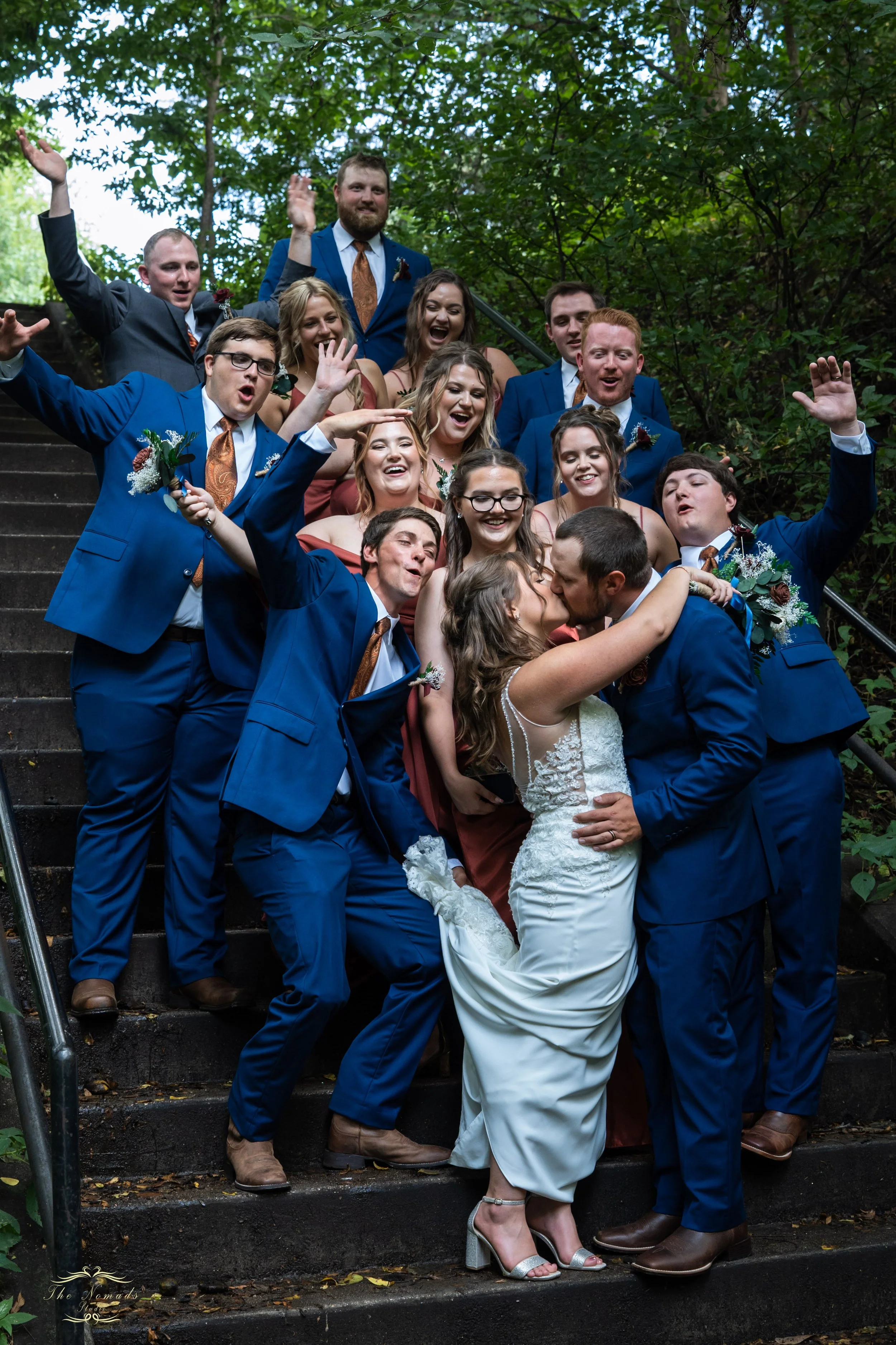 A group of wedding guests and the bride and groom on outdoor stairs, celebrating and kissing, surrounded by green trees.