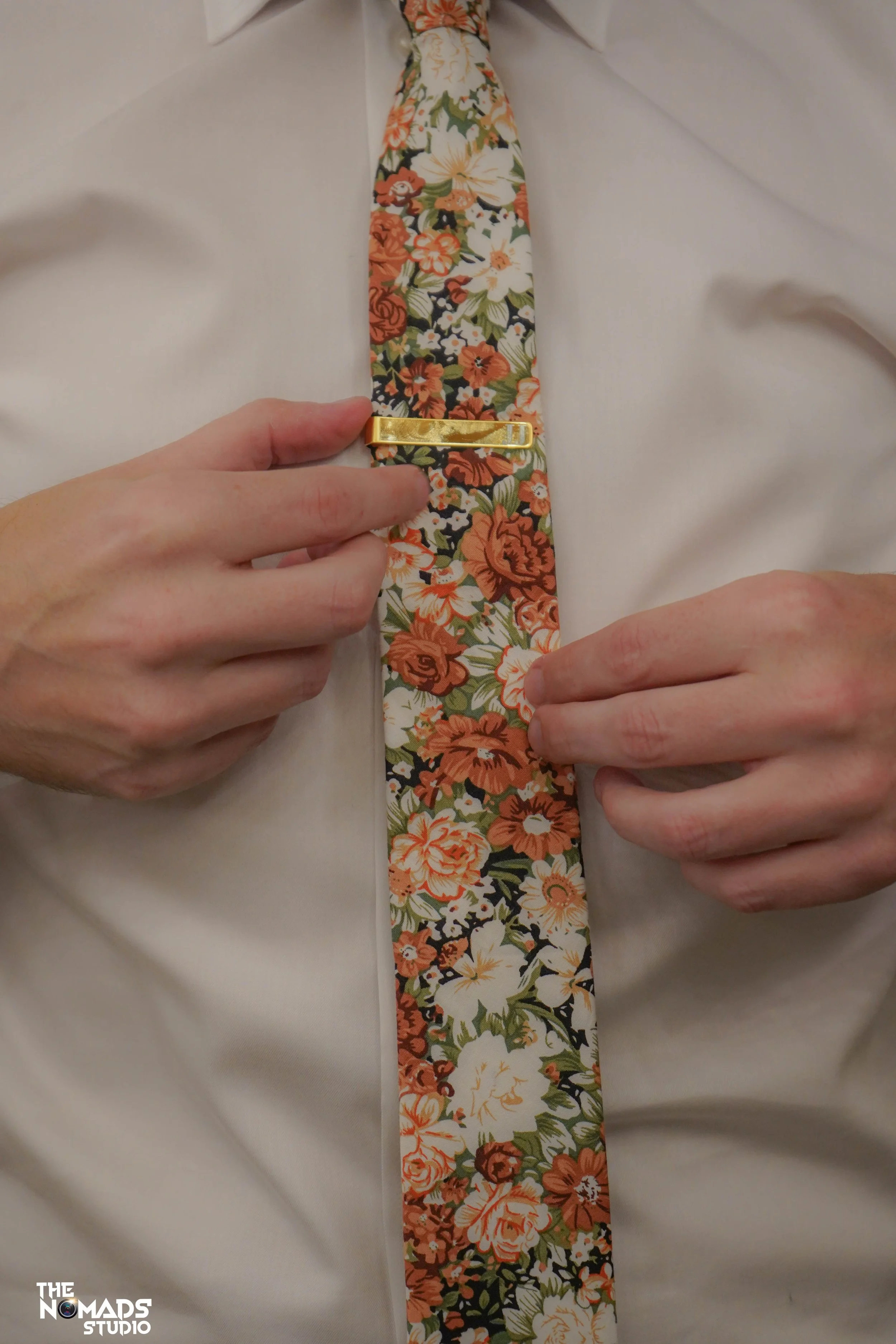 A close-up of a person wearing a white dress shirt with a floral tie featuring orange, white, and green colors, secured with a gold tie clip.