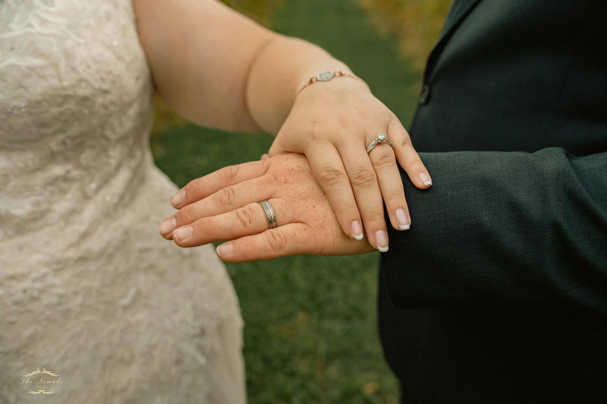 Close-up of a bride and groom holding hands, showing wedding rings, with the bride wearing a bracelet and a lace dress, outside in a grassy area.