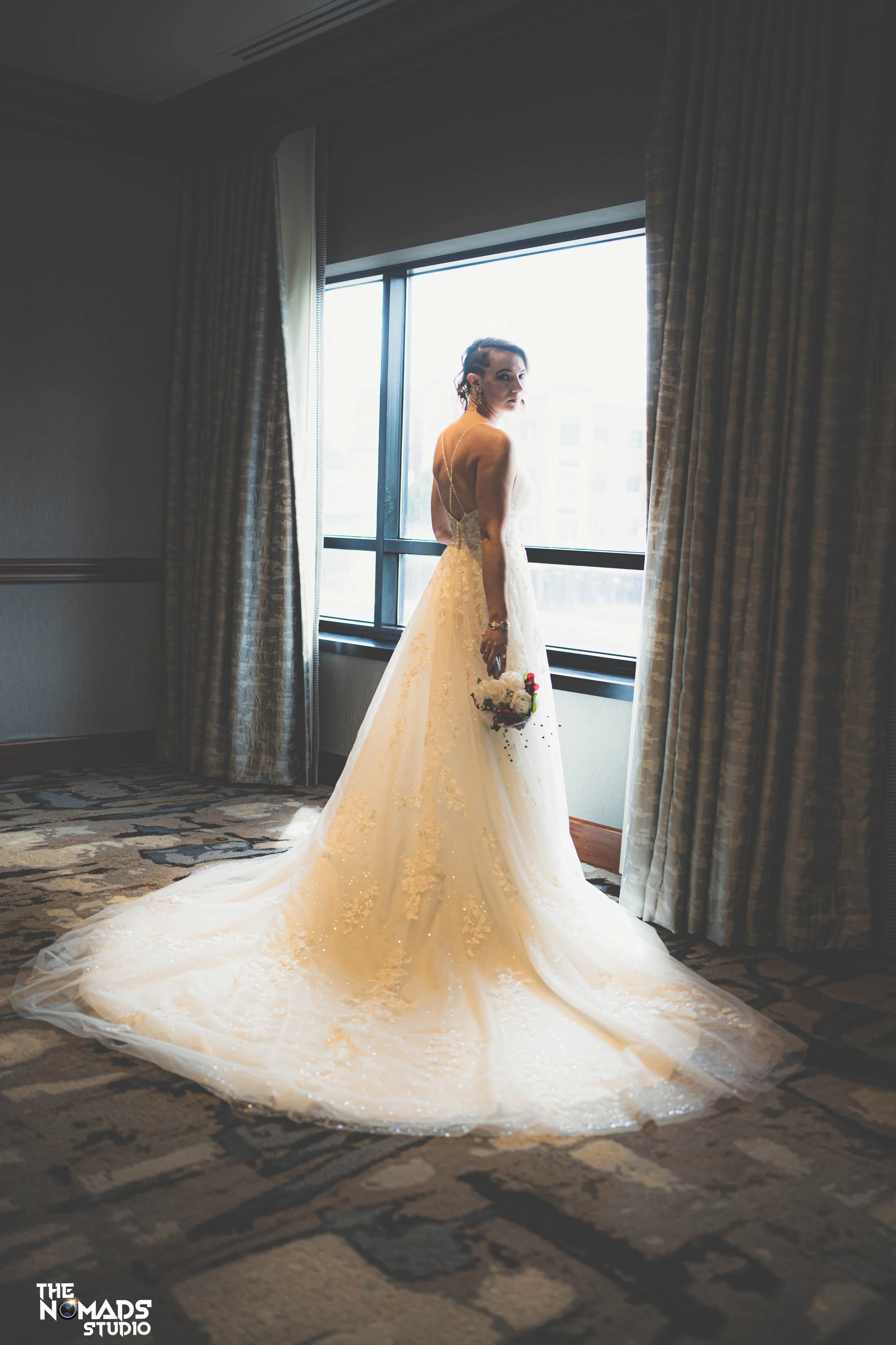 A bride in a white wedding gown with lace details stands by a large window, holding a bouquet of flowers, with curtains partially pulled back, in a hotel room.