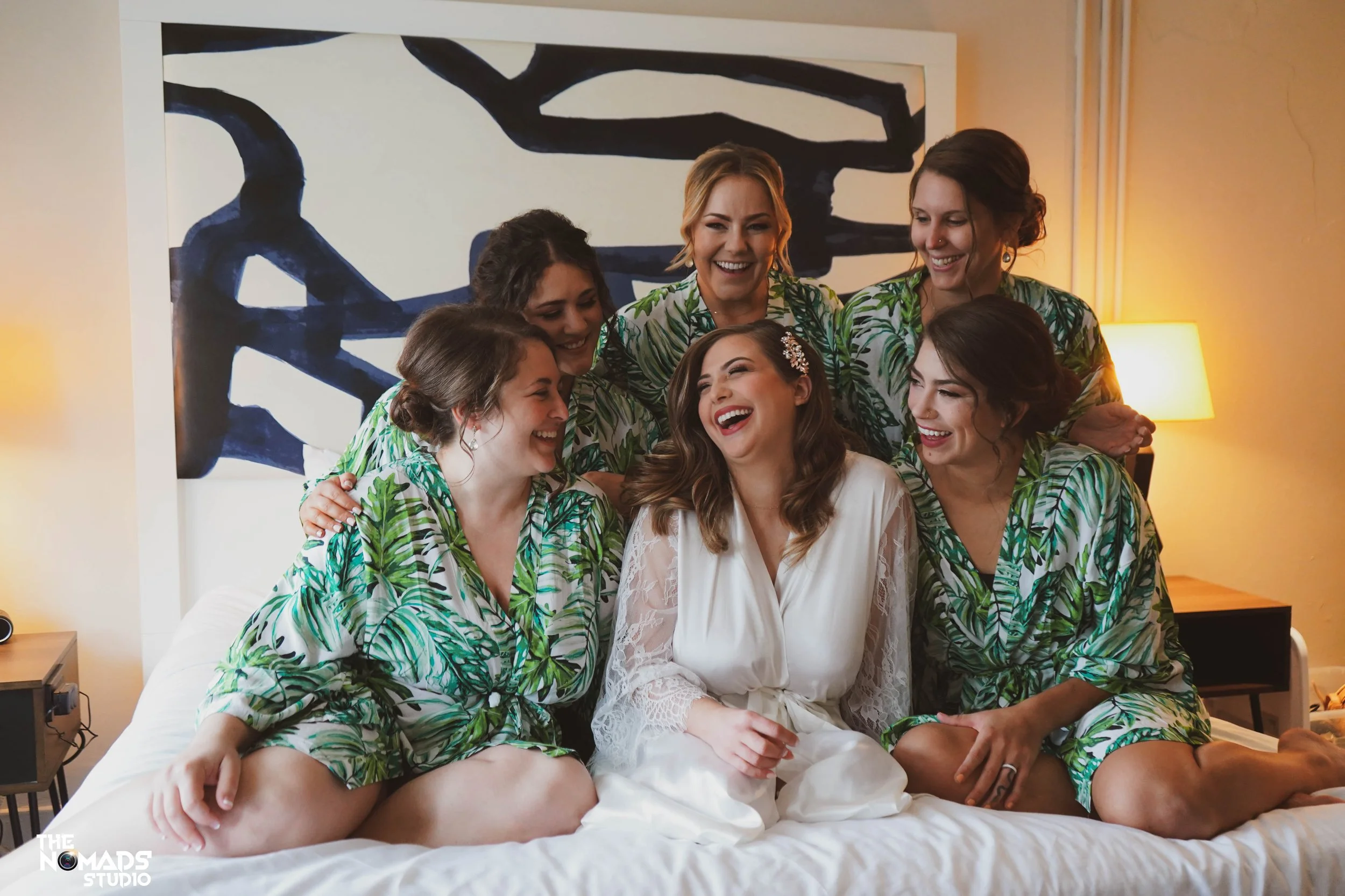 A group of seven women in matching tropical print robes gathered around a woman in a white robe, laughing and smiling in a warmly lit room with a large abstract black and white painting behind them and a small lamp to the right.