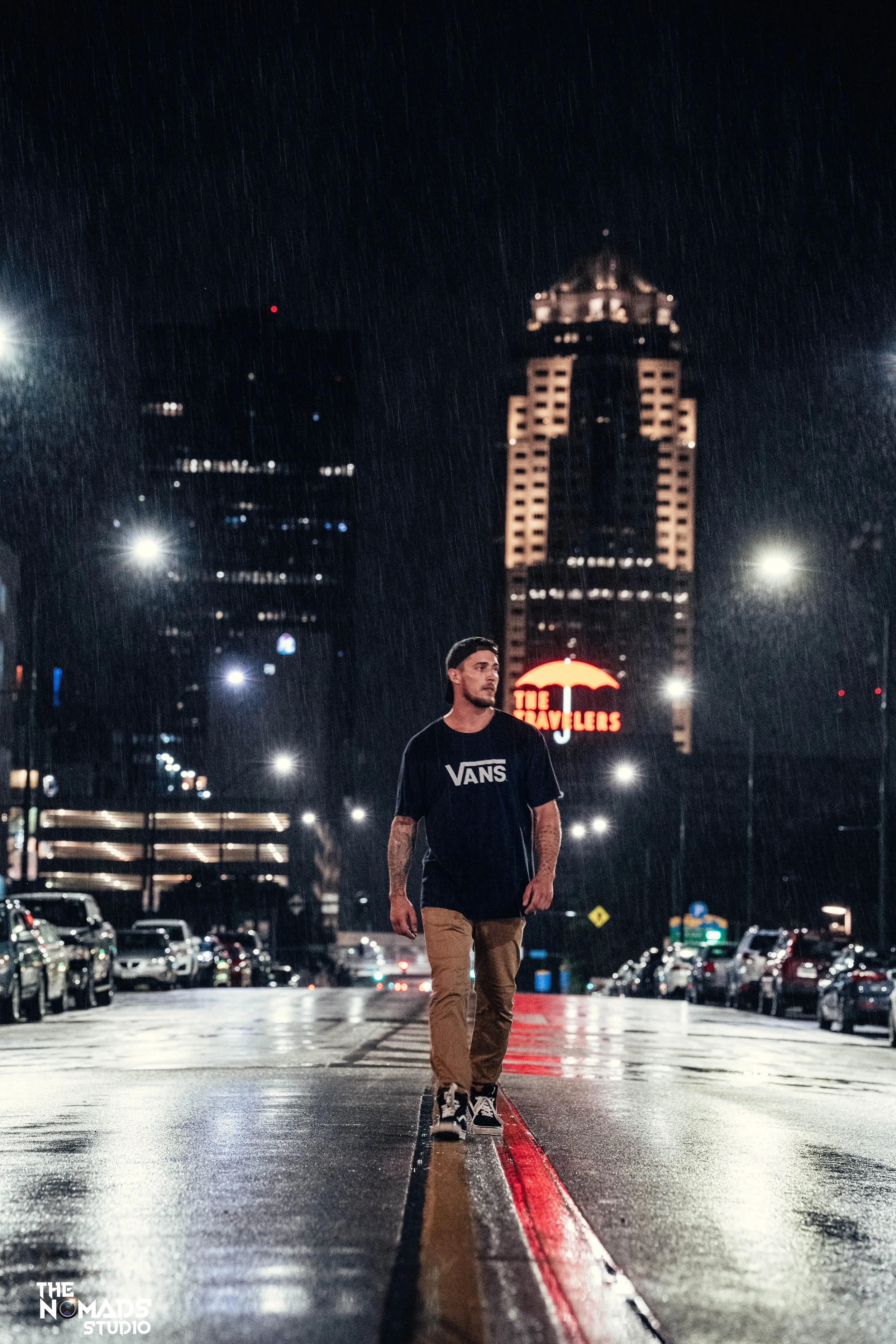 A young man with tattoos wearing a black Vans t-shirt and beige pants walking on a wet city street at night, with tall buildings illuminated in the background and a neon sign that reads 'The Travelers.'