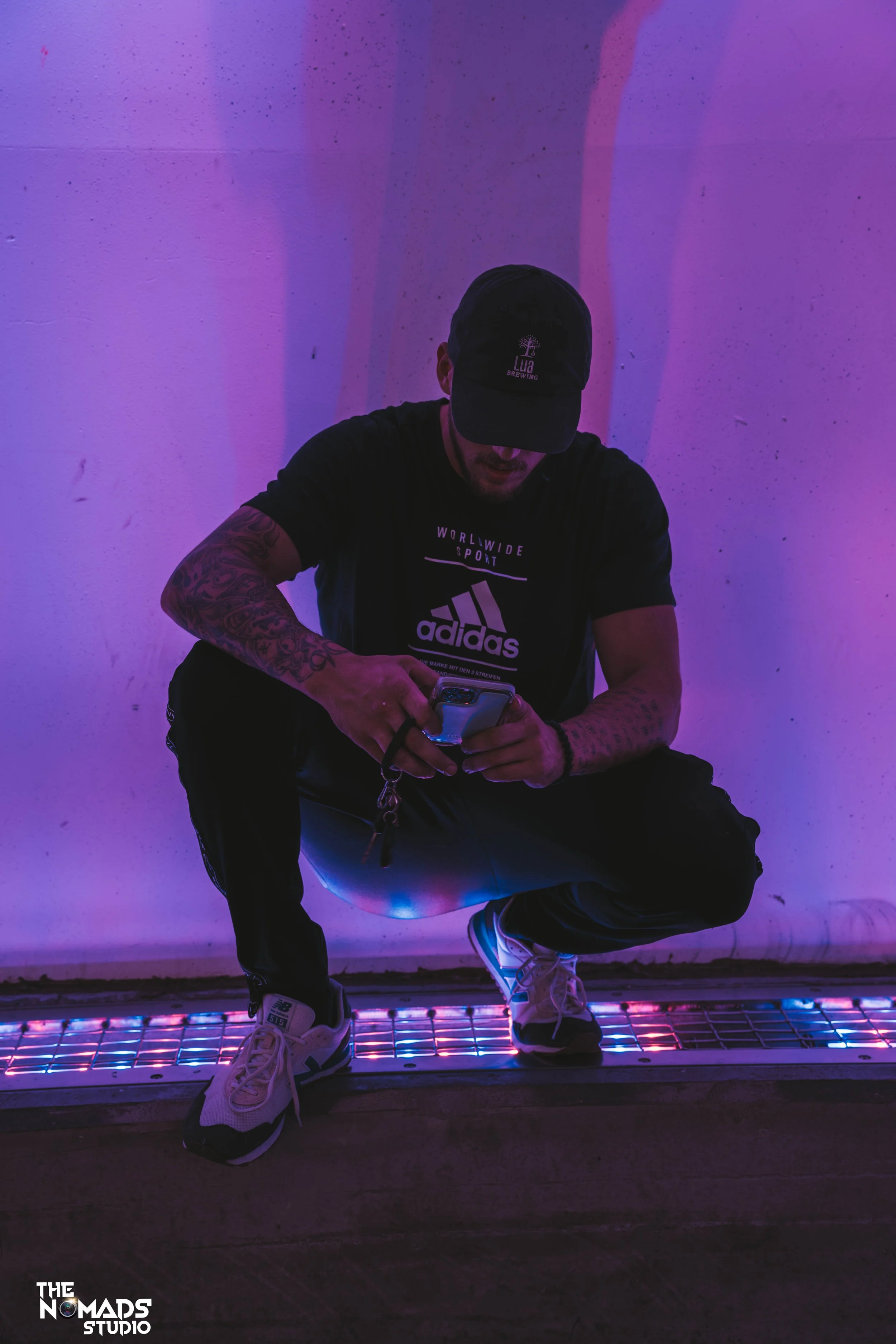 A man with tattoos sitting on a metallic bench, wearing a black cap and black T-shirt, looking at his phone. The background is illuminated with purple and pink lights, creating a colorful atmosphere.