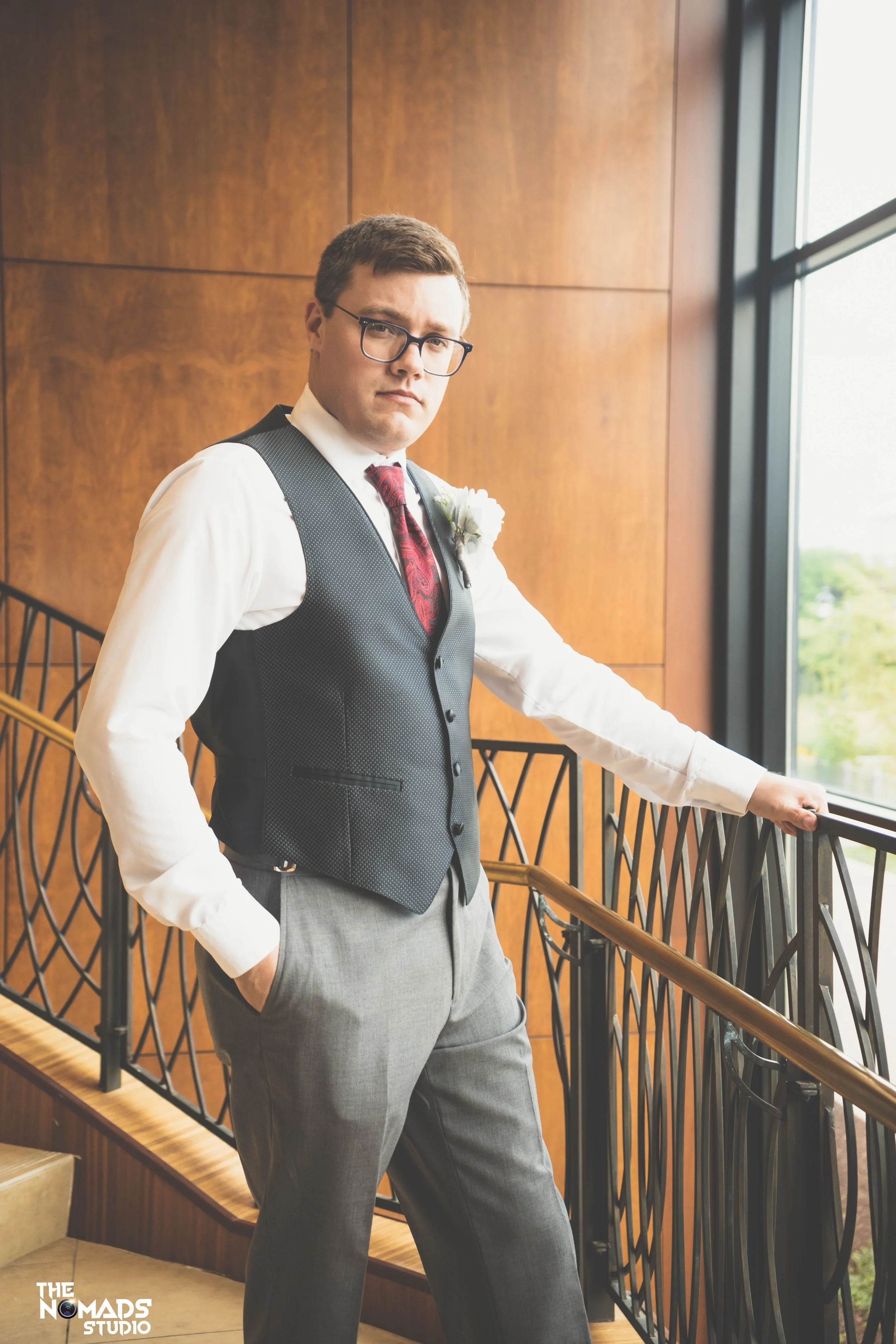 A man in a gray pants, white shirt, black vest, and red patterned tie standing by a window with a view of green trees, holding onto a black railing in an indoor setting with wooden walls.