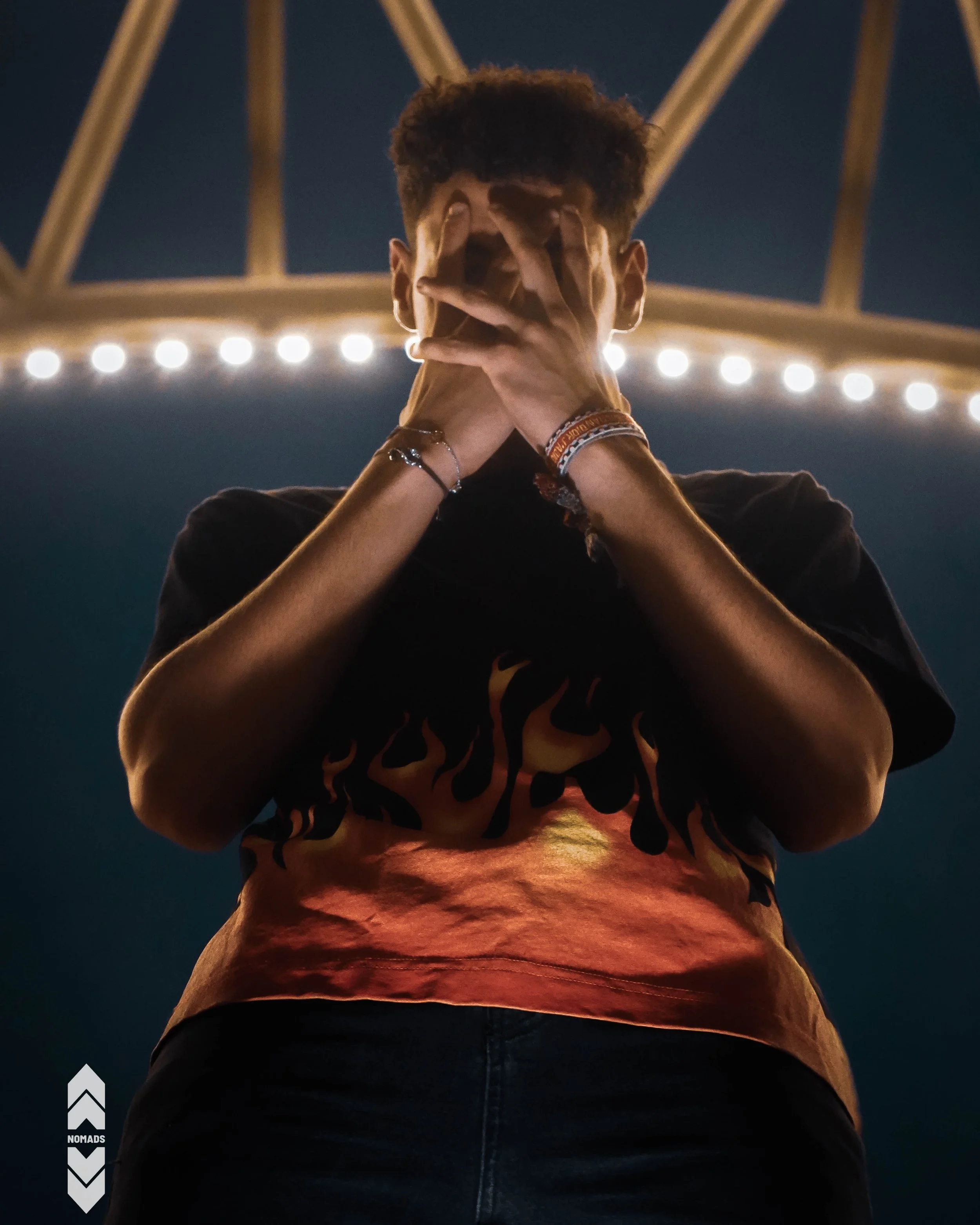 A person standing with hands covering their face, wearing a black shirt with orange flames and bracelets, against a backdrop of a bridge with lights at night.
