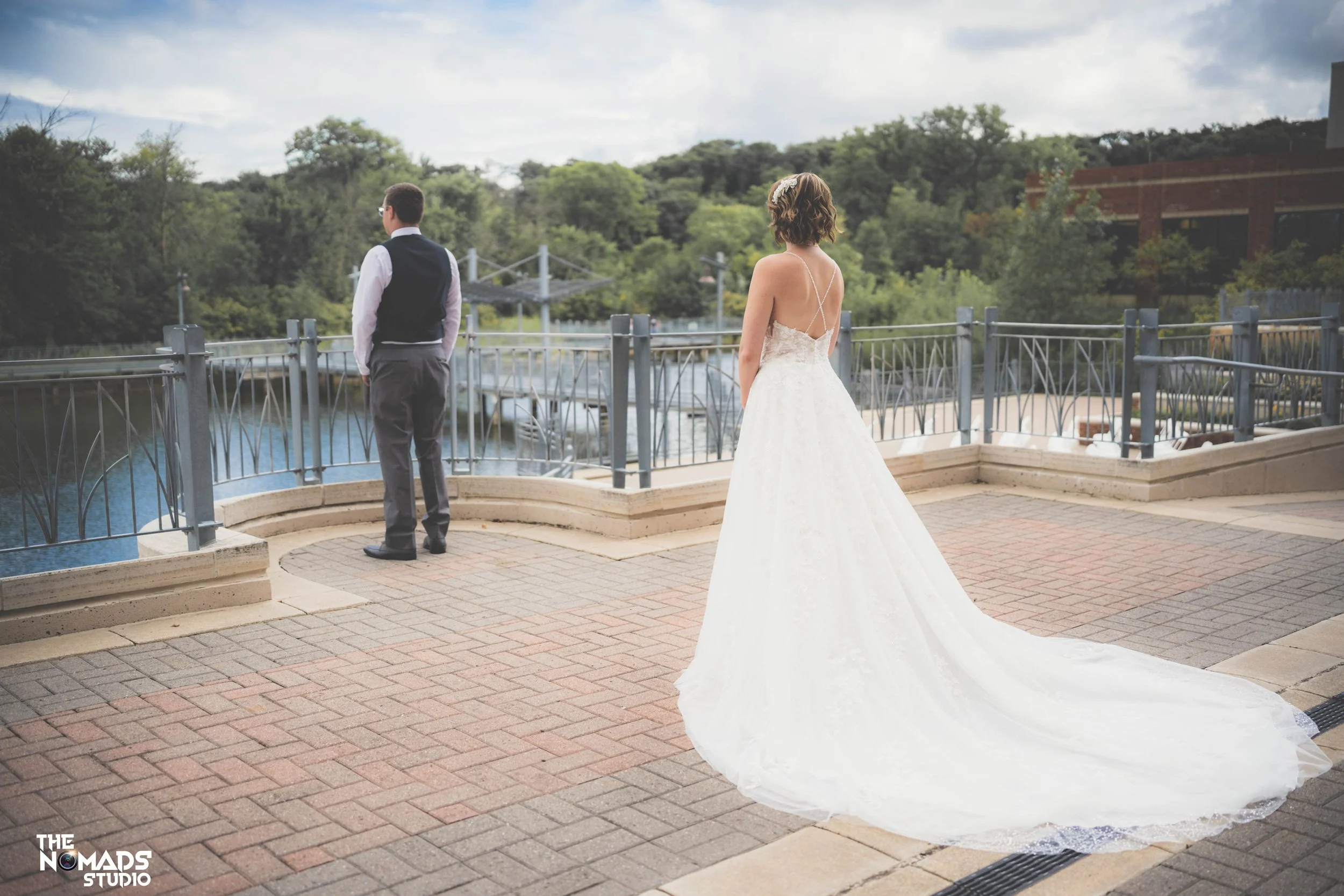 A bride in a white wedding gown with a train stands on a brick patio, watching a groom in a black vest and gray pants near a body of water, with trees and a cloudy sky in the background.