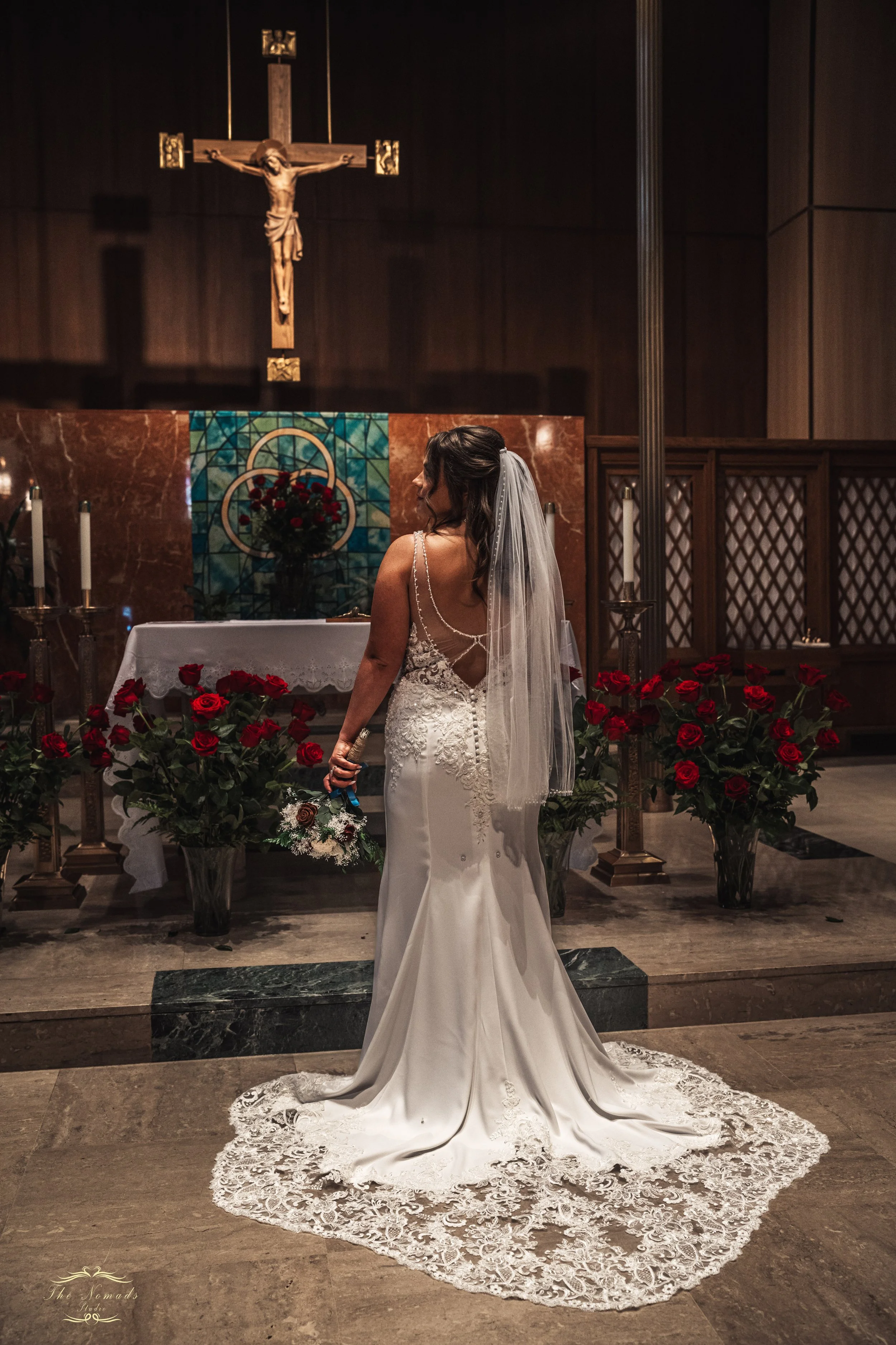 A bride in a white wedding dress with lace detailing stands in front of an altar decorated with red roses, holding a bouquet, in a church with a crucifix on the wall above.