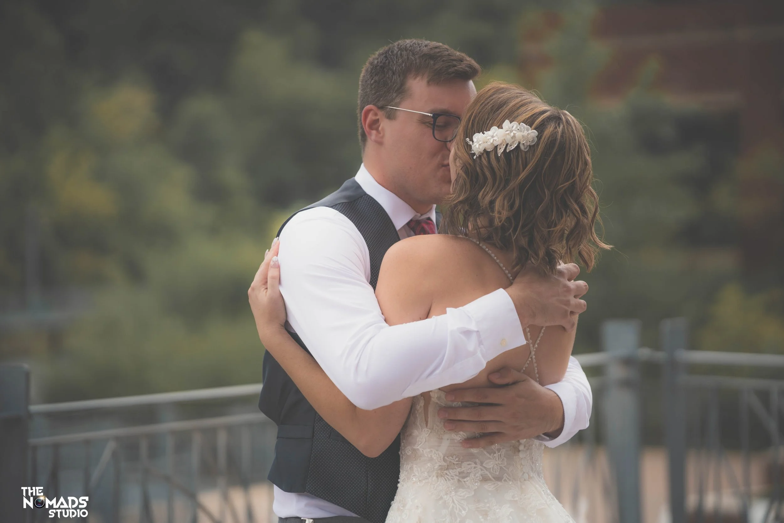 A couple, dressed in wedding attire, embracing outdoors on a cloudy day, with greenery and a railing in the background.
