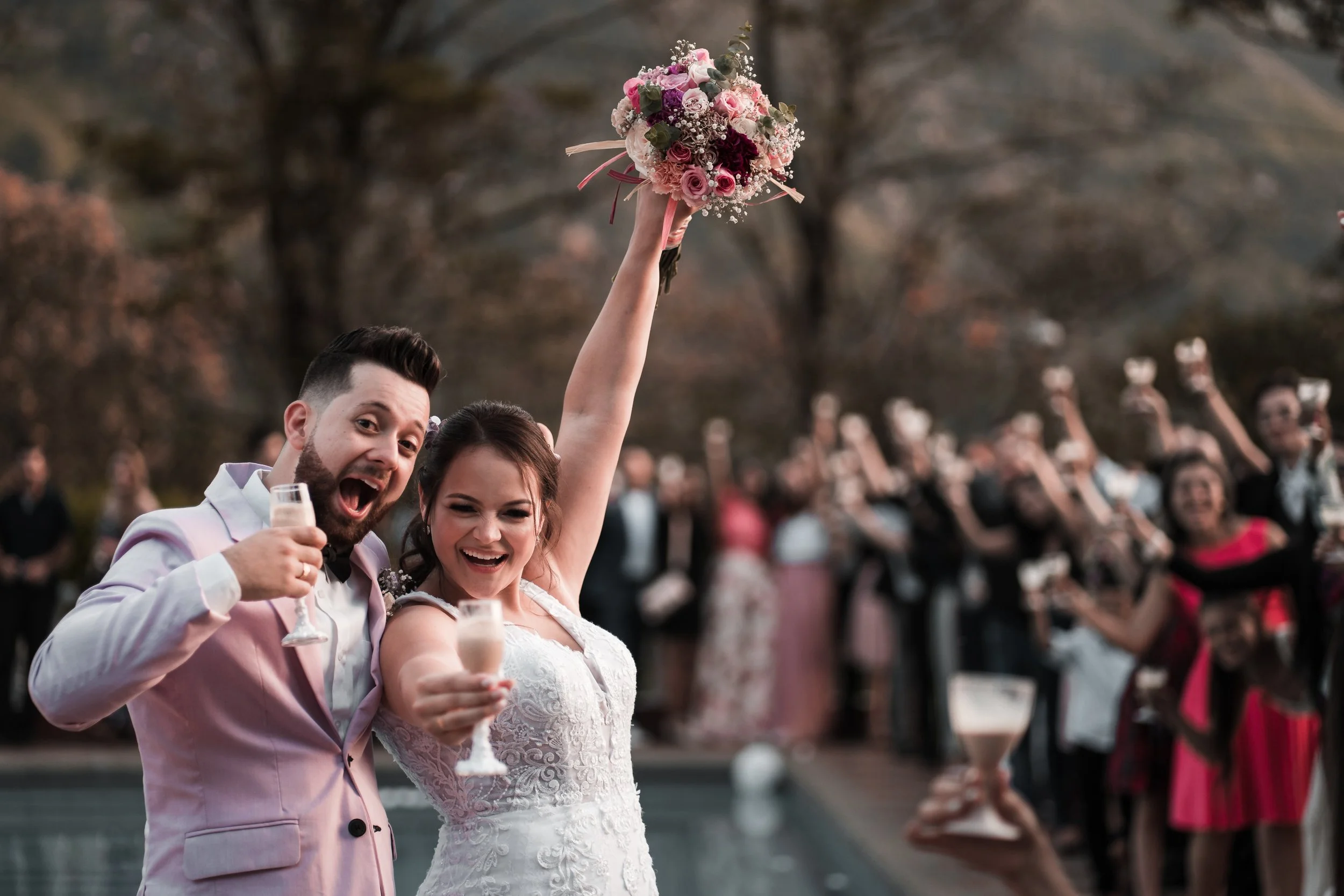A newlywed couple at a celebration outdoors. The groom in a pink suit holds a glass of champagne and smiles. The bride holds a bouquet of pink and white flowers above her head, smiling. Guests in the background are raising glasses and appear to be ce