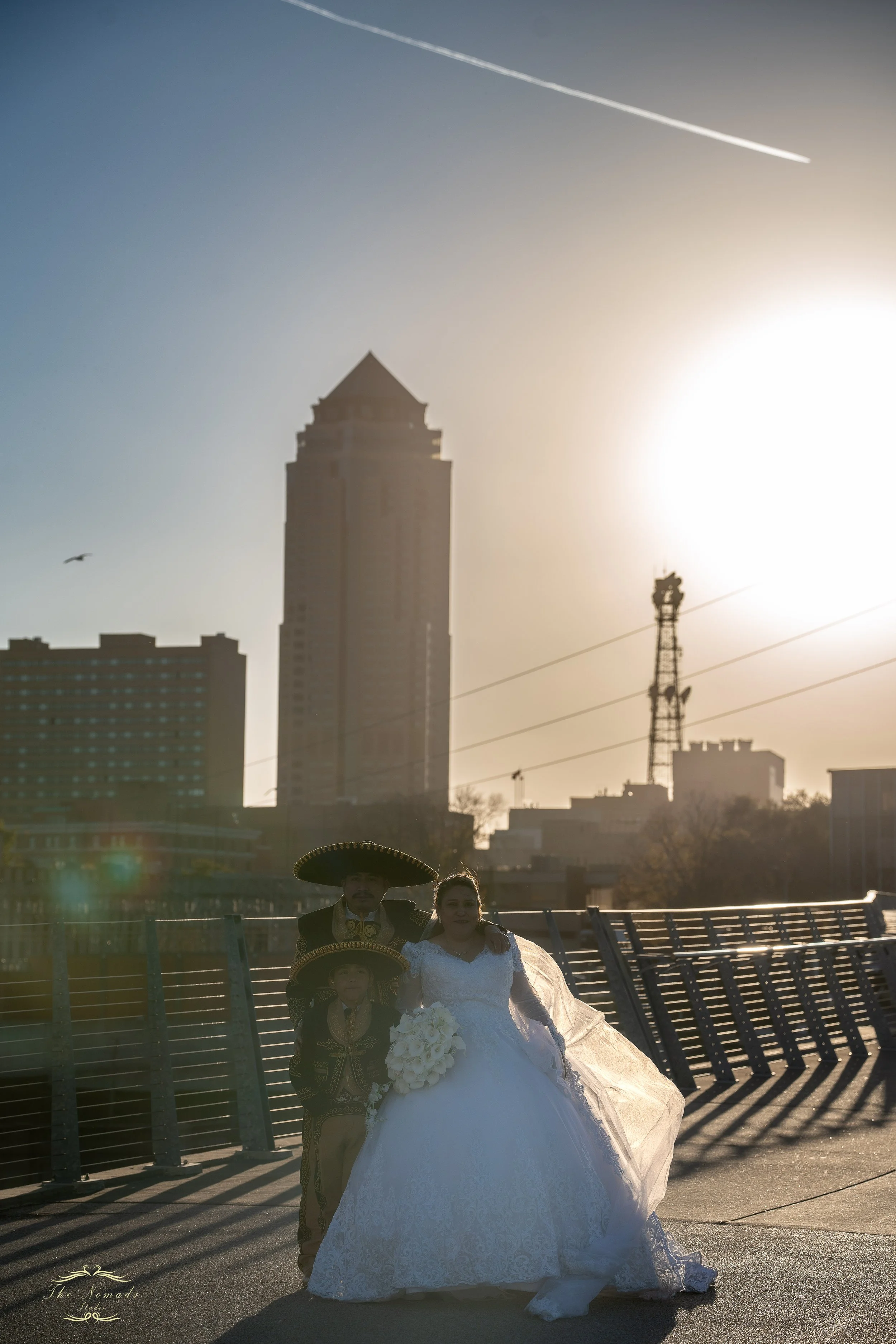 A bride in a white wedding dress standing outdoors with a man dressed in traditional Mexican attire and a child in front of her, with a city skyline and a bright sun in the background.