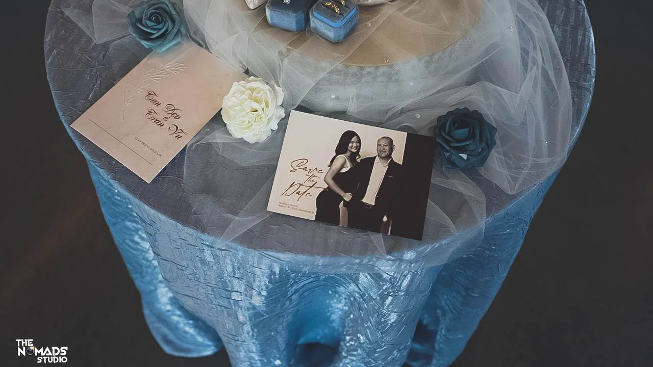 A wedding or engagement display table with a photo of a couple, decorative flowers, and a card with calligraphy script, draped with sheer fabric and a textured blue tablecloth.