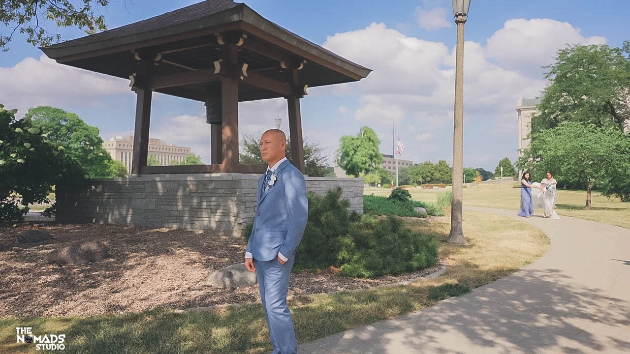 A man in a light blue suit standing near a gazebo in a park, with three women on the path in the background, two of whom are wearing long dresses, and an American flag is visible in the distance under a partly cloudy sky.