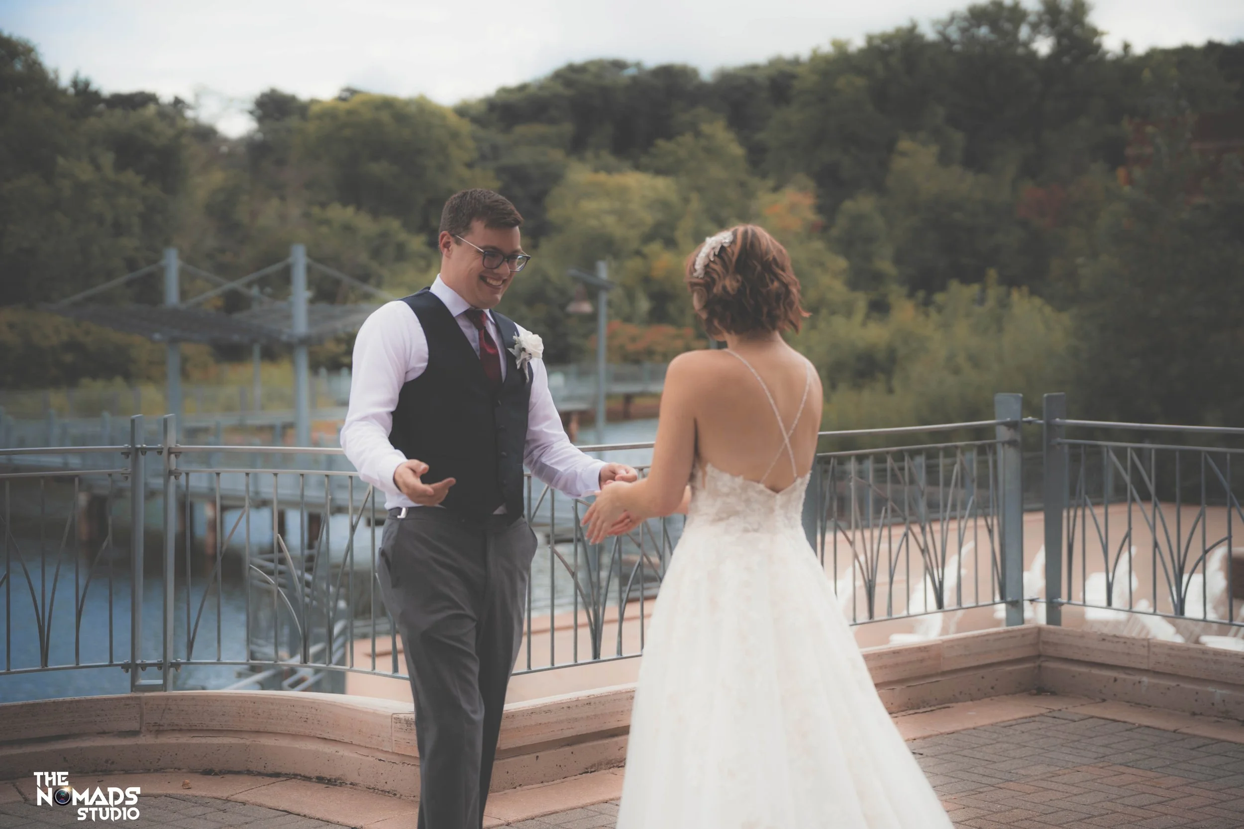 A bride and groom holding hands and smiling at each other on an outdoor terrace with a view of trees and a bridge in the background.