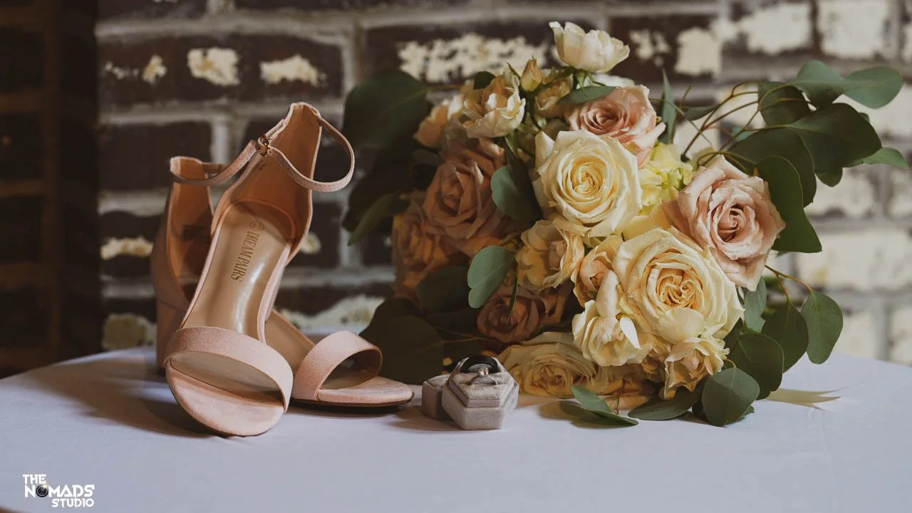 A pair of peach high heel sandals, a bouquet of cream and blush roses with greenery, and a small jewelry box with rings on a white tablecloth against a brick wall background.