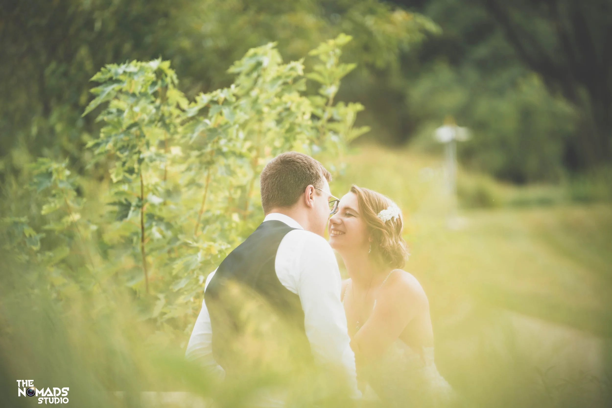 A couple, possibly newlyweds, in wedding attire sharing an intimate moment outdoors among lush green foliage with soft sunlight.