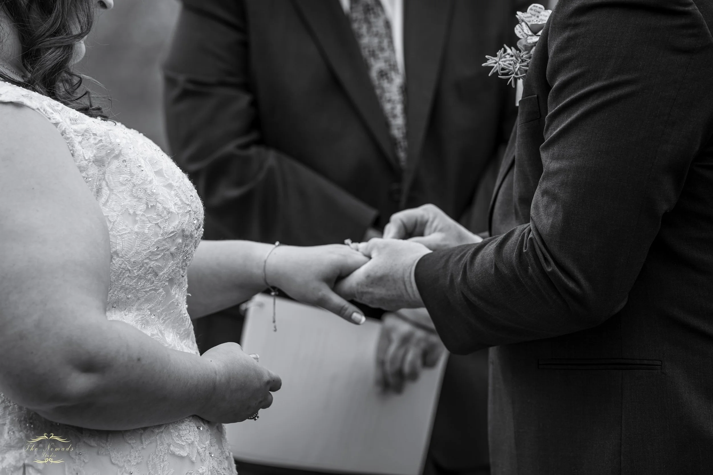 A bride and groom exchange vows during a wedding ceremony. The bride is wearing a lace wedding dress, and the groom is wearing a dark suit with a boutonniere. They are holding hands, and an officiant is standing between them.