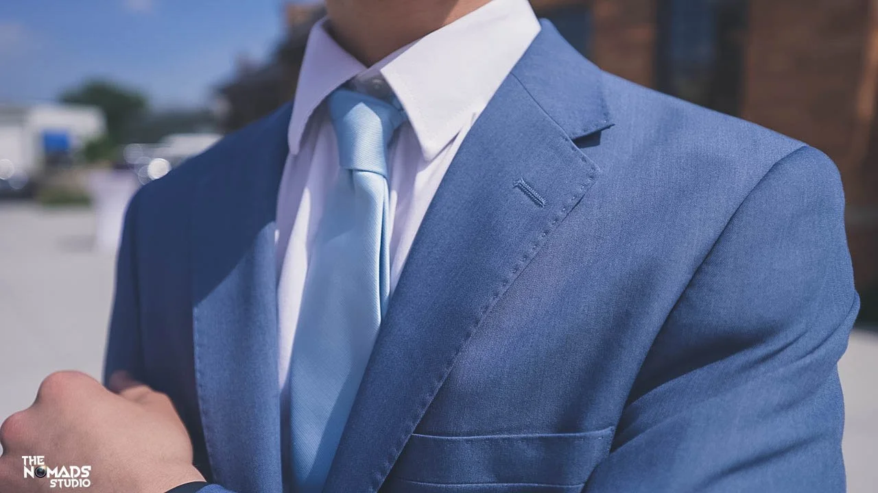 Close-up of a man wearing a blue suit, white shirt, and light blue tie, standing outside with blurred background