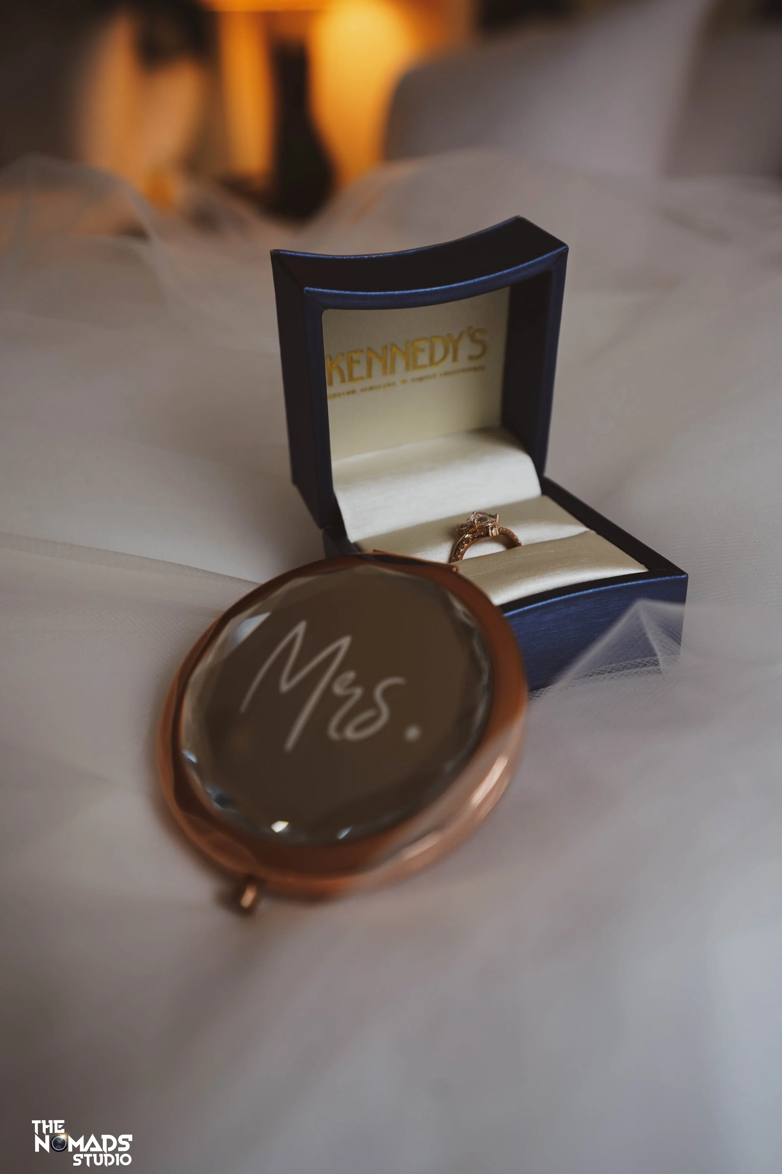 An open black ring box with gold lettering inside reads 'KENNEDY'S' and contains a gold ring with a large gemstone. In front of the box is a round mirror with a rose gold border. The background is blurred with warm lighting.