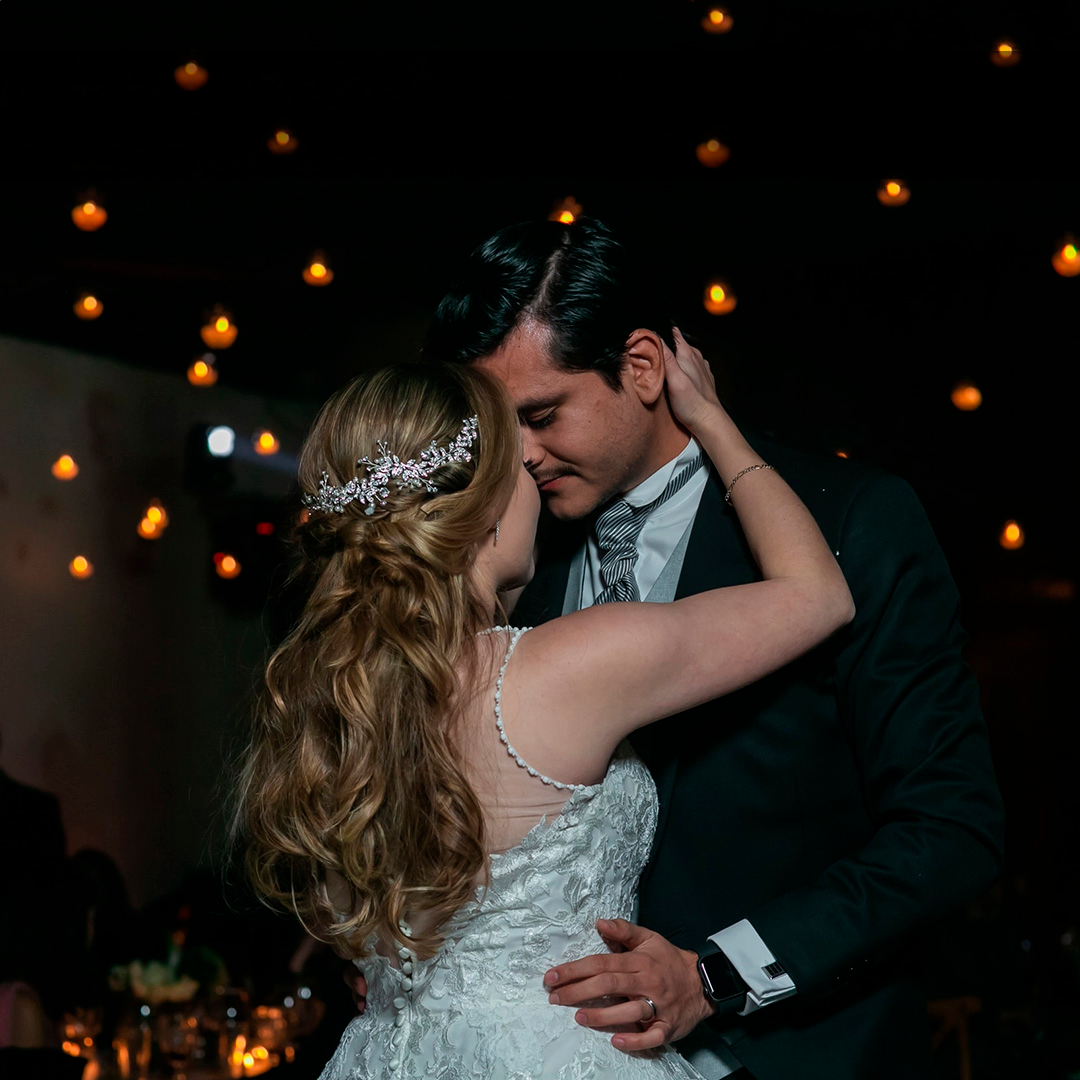 A bride and groom dancing closely at their wedding reception, with warm string lights overhead and guests seated in the background.