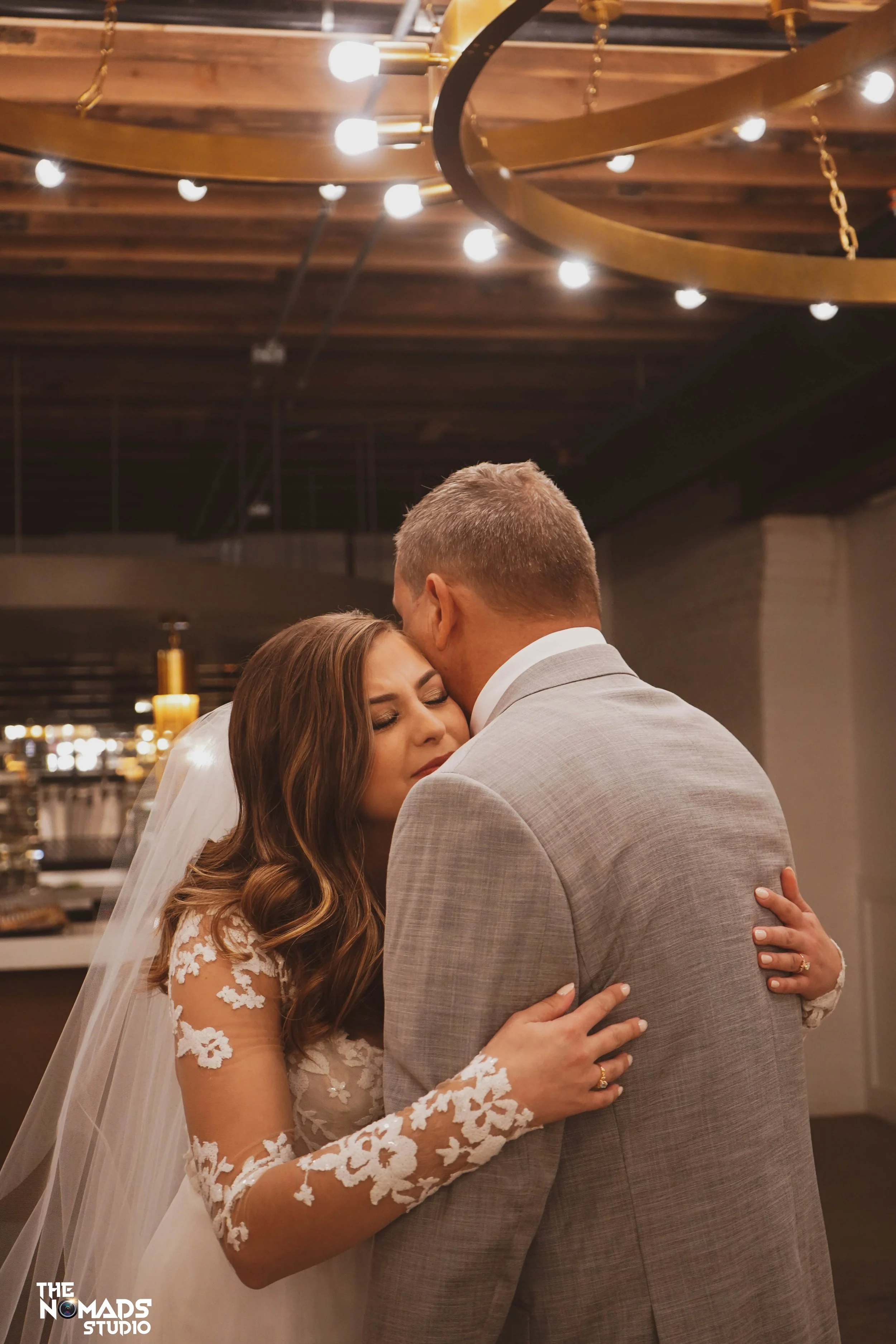 A bride and groom share an intimate moment during their wedding ceremony, with the bride in a lace wedding dress and the groom in a gray suit, embracing in a warmly lit venue with circular lighting fixtures overhead.