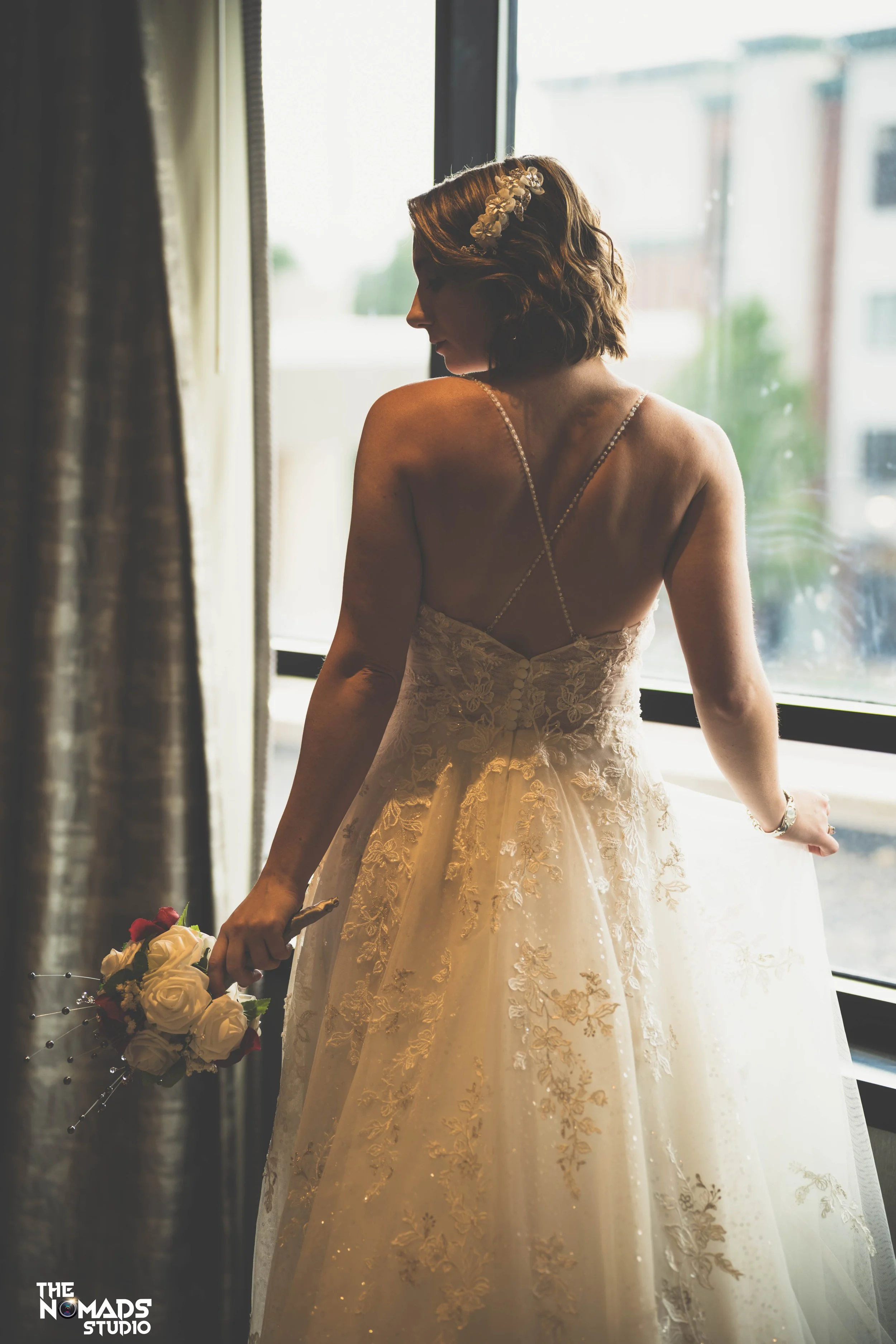 A bride in a lace wedding dress with spaghetti straps stands by a window, holding a bouquet of white and red roses, looking away. She wears a floral hairpiece, and sunlight streams through the window, illuminating her silhouette.