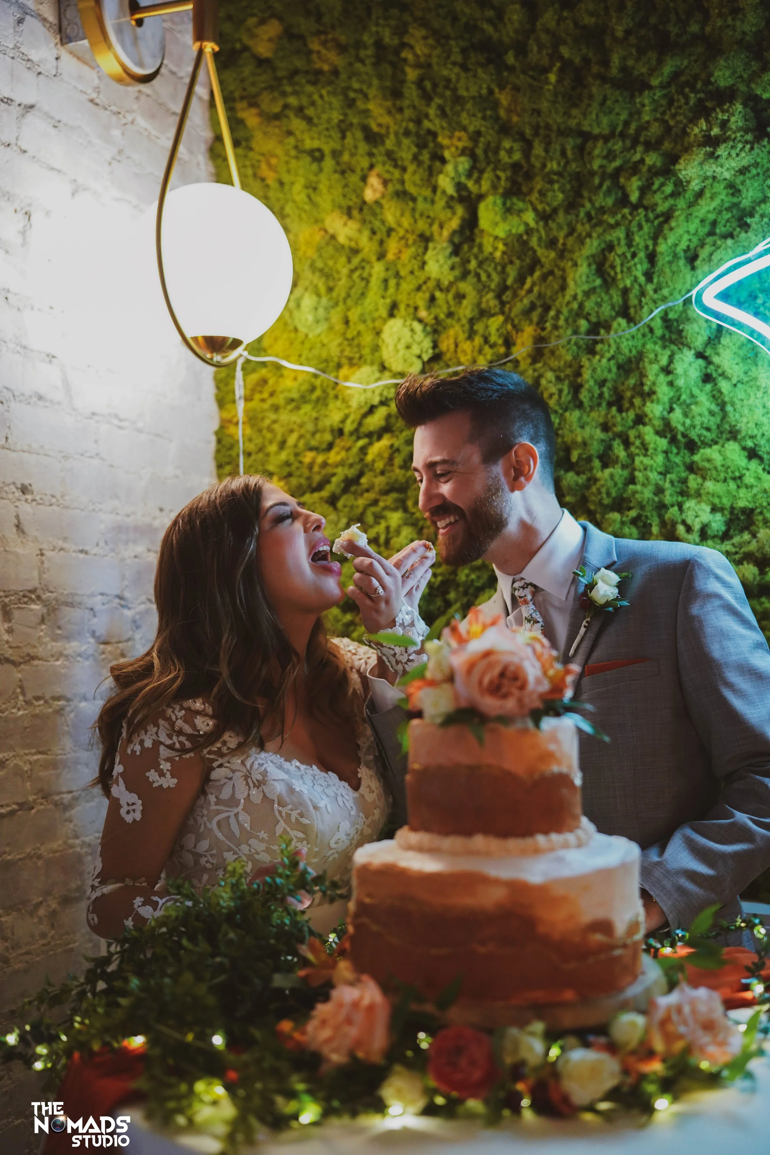 A newlywed couple sharing a cake at their wedding reception, with a decorative greenery wall and a neon light in the background.