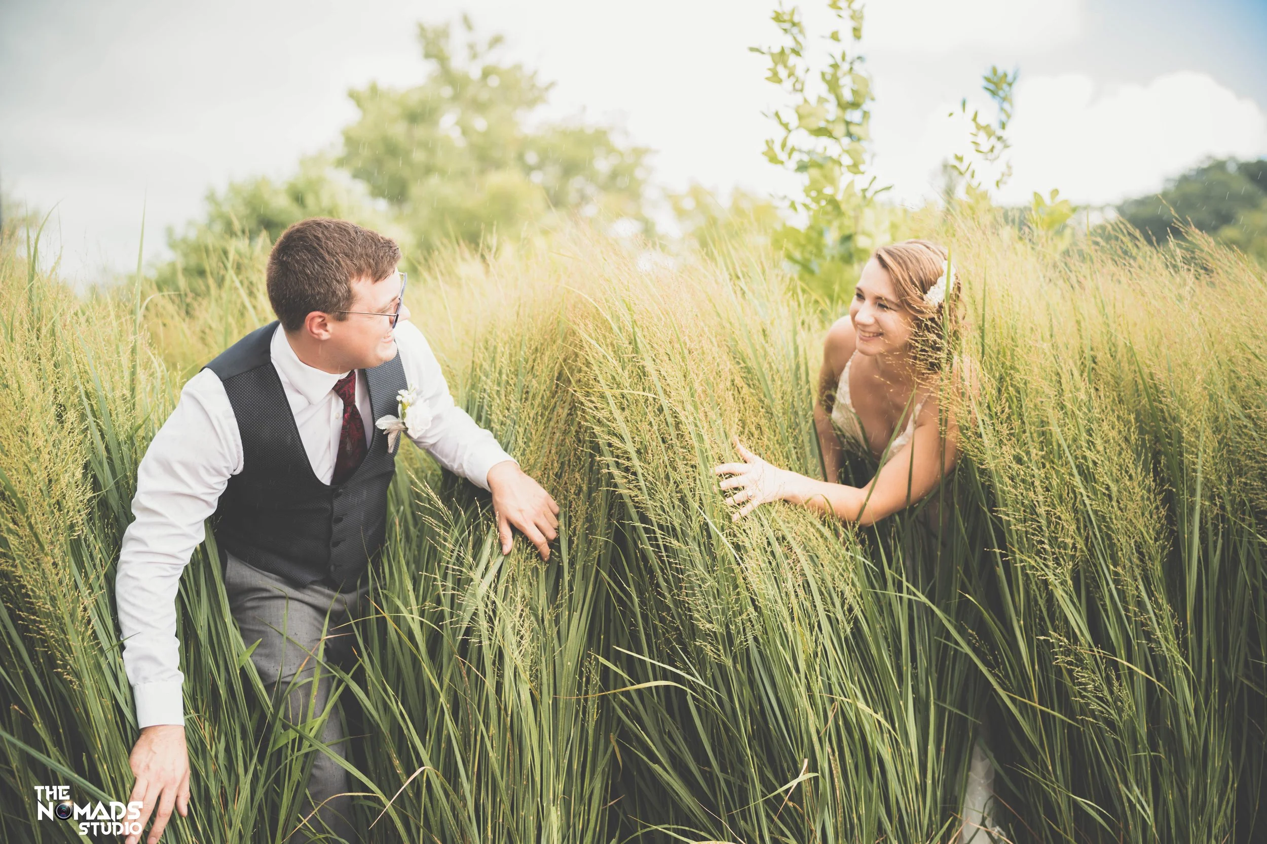 A man and a woman smiling at each other while lying in a field of tall grass and yellow wildflowers outdoors on a sunny day.