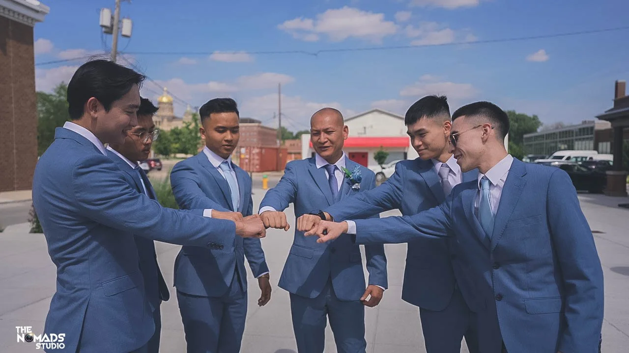 Six men in blue suits gathering outside, touching fists together in a gesture of friendship or unity, with a bright sky and buildings in the background.
