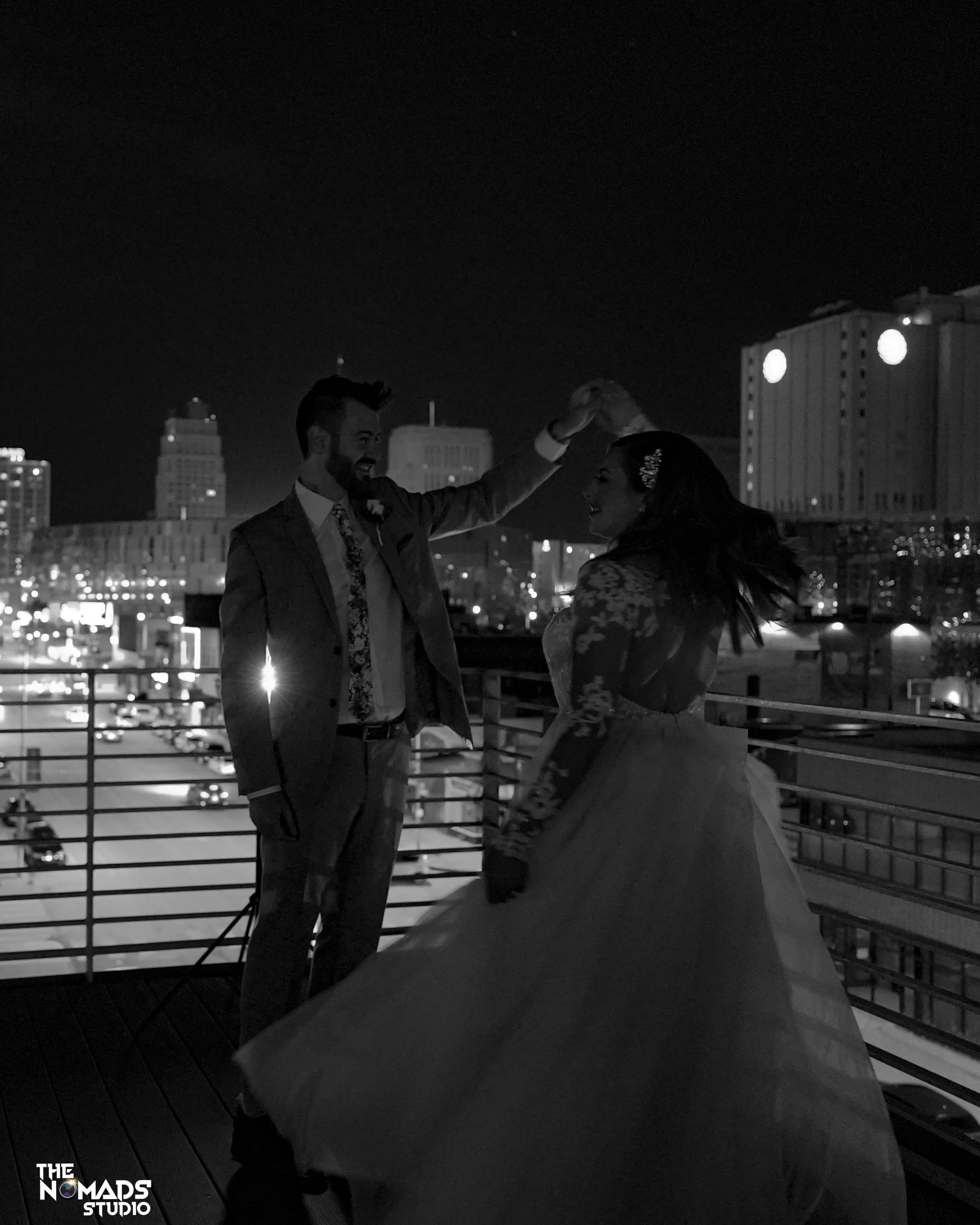 A couple dancing on a rooftop at night with city buildings in the background. The man is in a suit and the woman in a wedding dress. They appear to be happy.