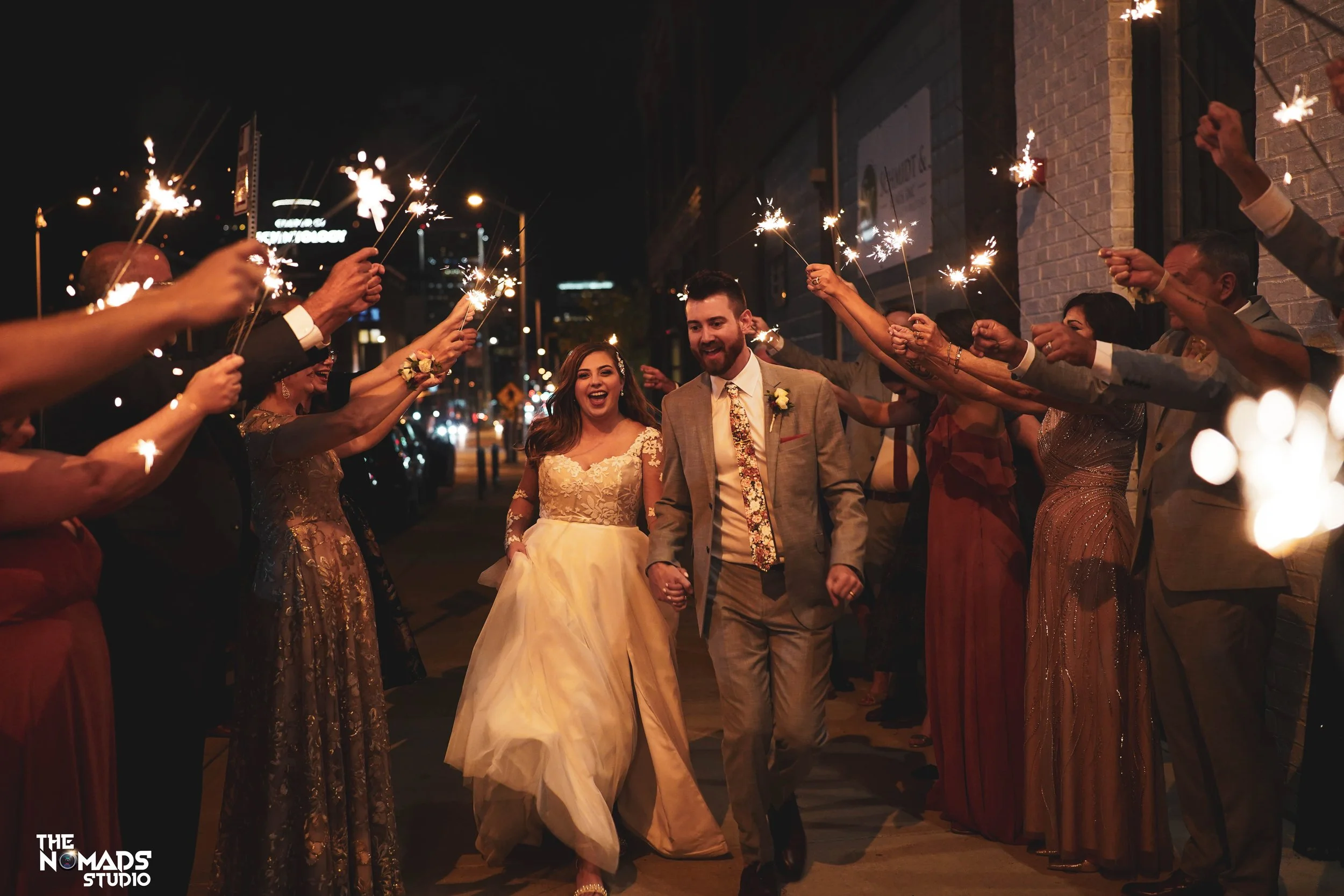 A newlywed couple walking hand in hand through a crowd of people holding sparklers outdoors at night to celebrate their wedding.