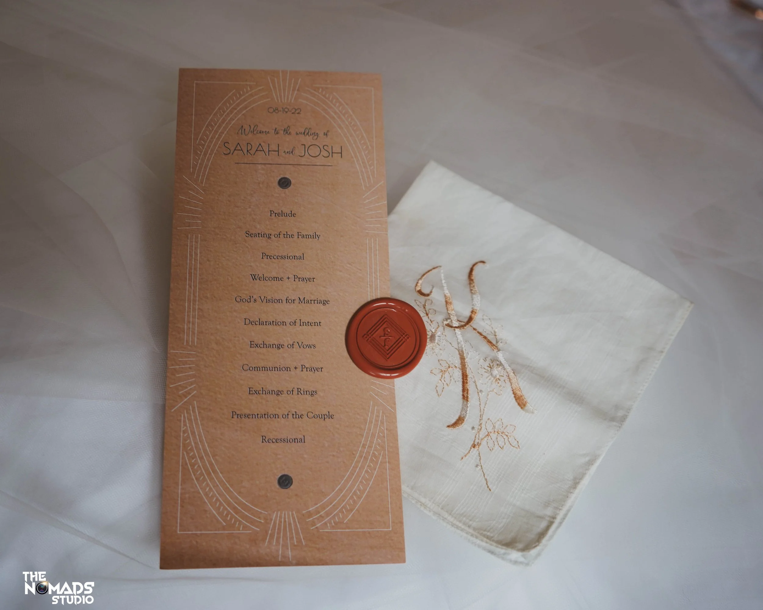 Wedding program or invitation card for Sarah and Josh's wedding, with details of the ceremony schedule, sealed with a red wax stamp, and white embroidered fabric nearby.