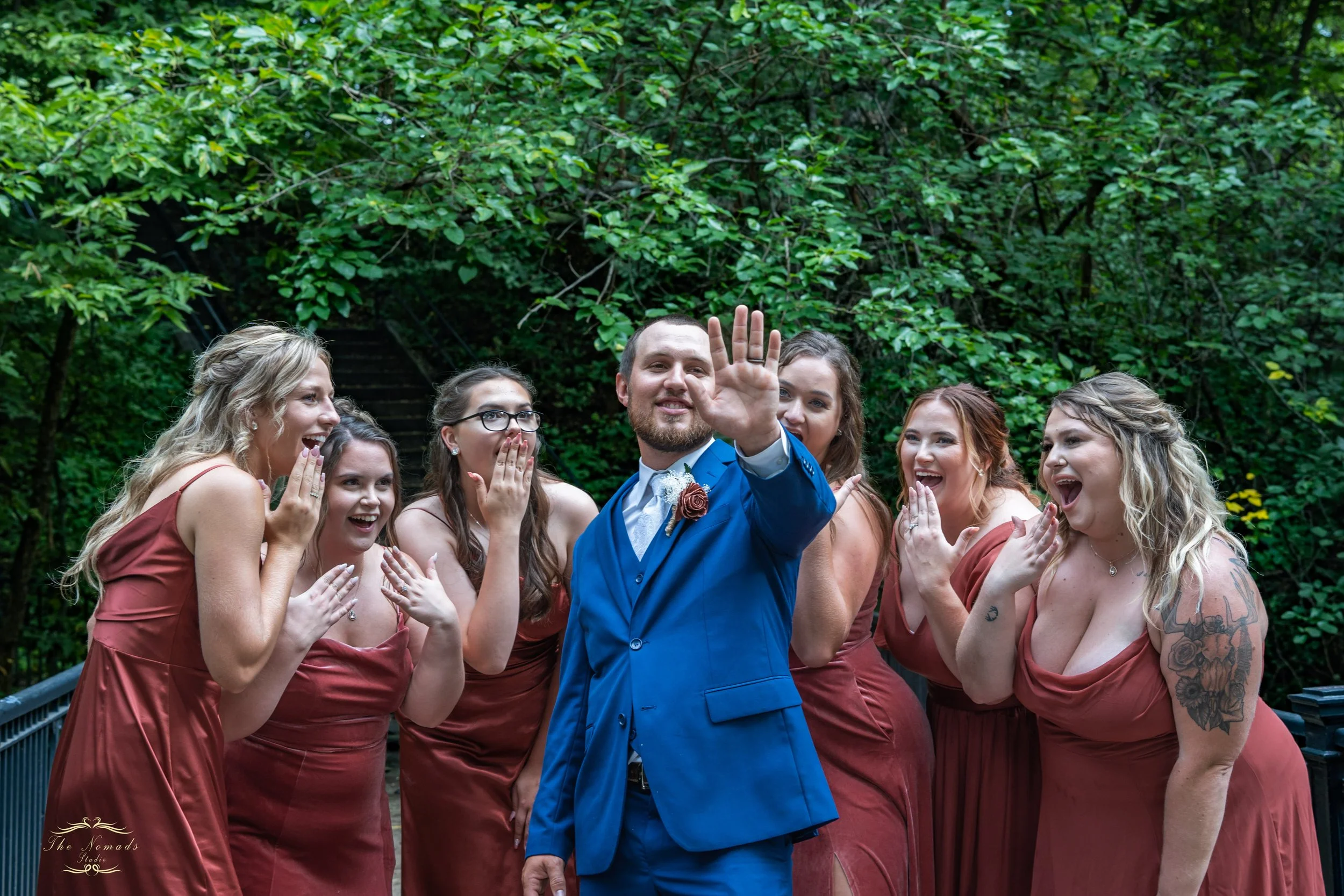 A man in a blue suit waving, surrounded by women in matching rust-colored dresses, outdoors with green trees in the background.