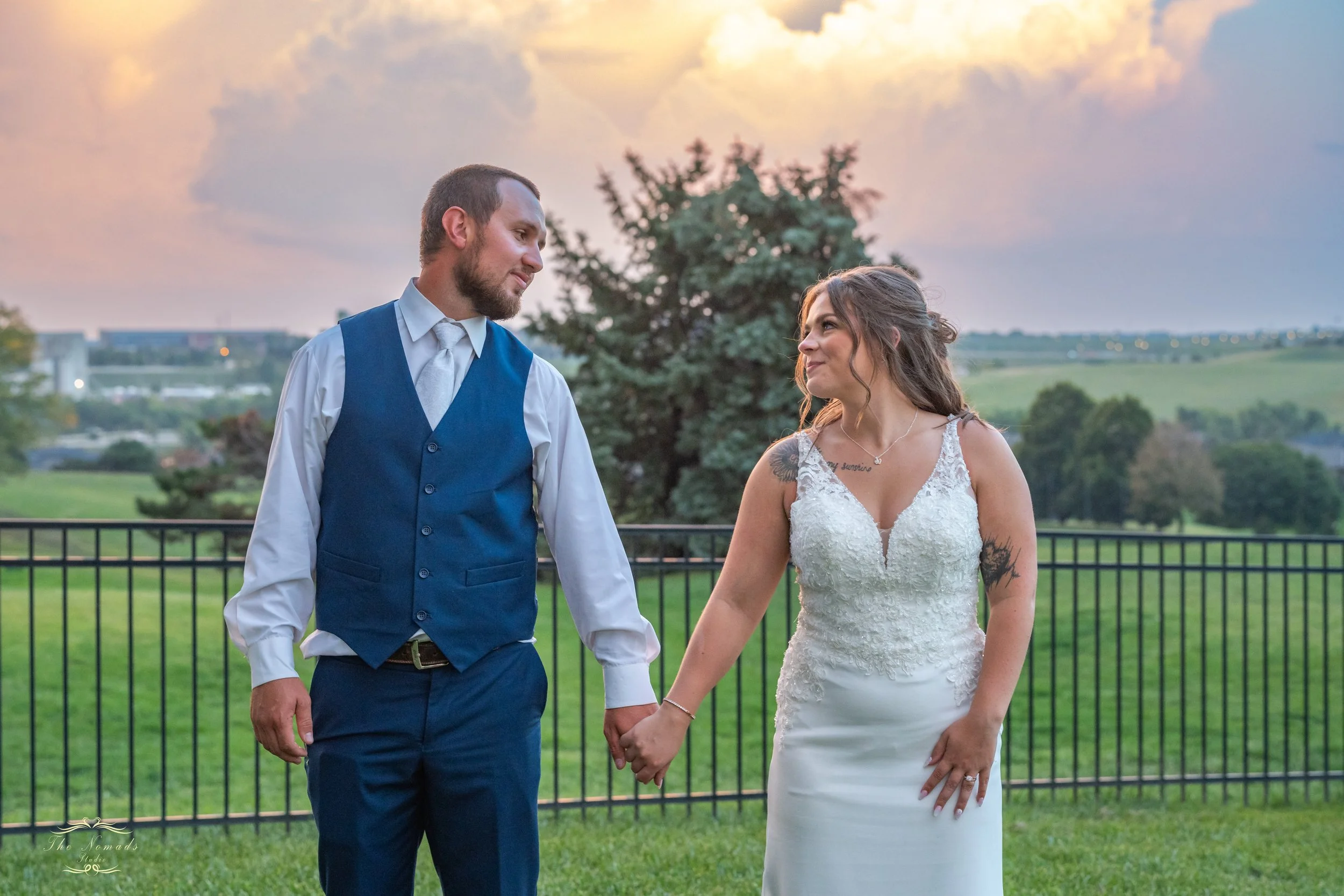 A couple, holding hands, is standing outdoors in formal wear, with a scenic background of trees and a cloudy sky during sunset.